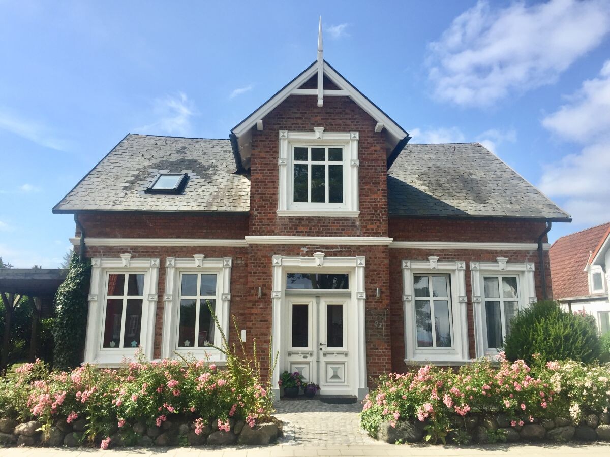 Rotes Backsteinhaus mit weißen Fensterrahmen, blauer Himmel, rosa Rosen im Vorgarten.