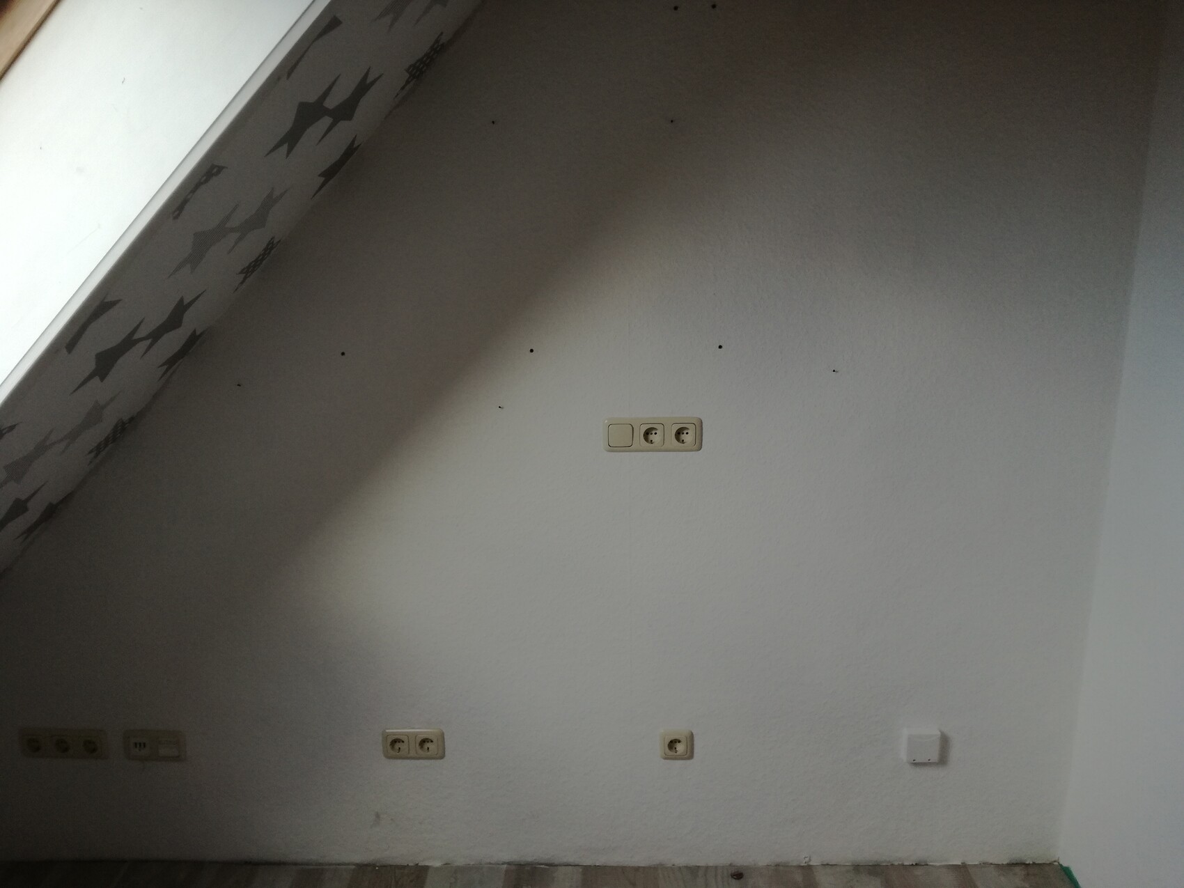 Graue Wand mit mehreren Steckdosen; links eine gemusterte Fläche.