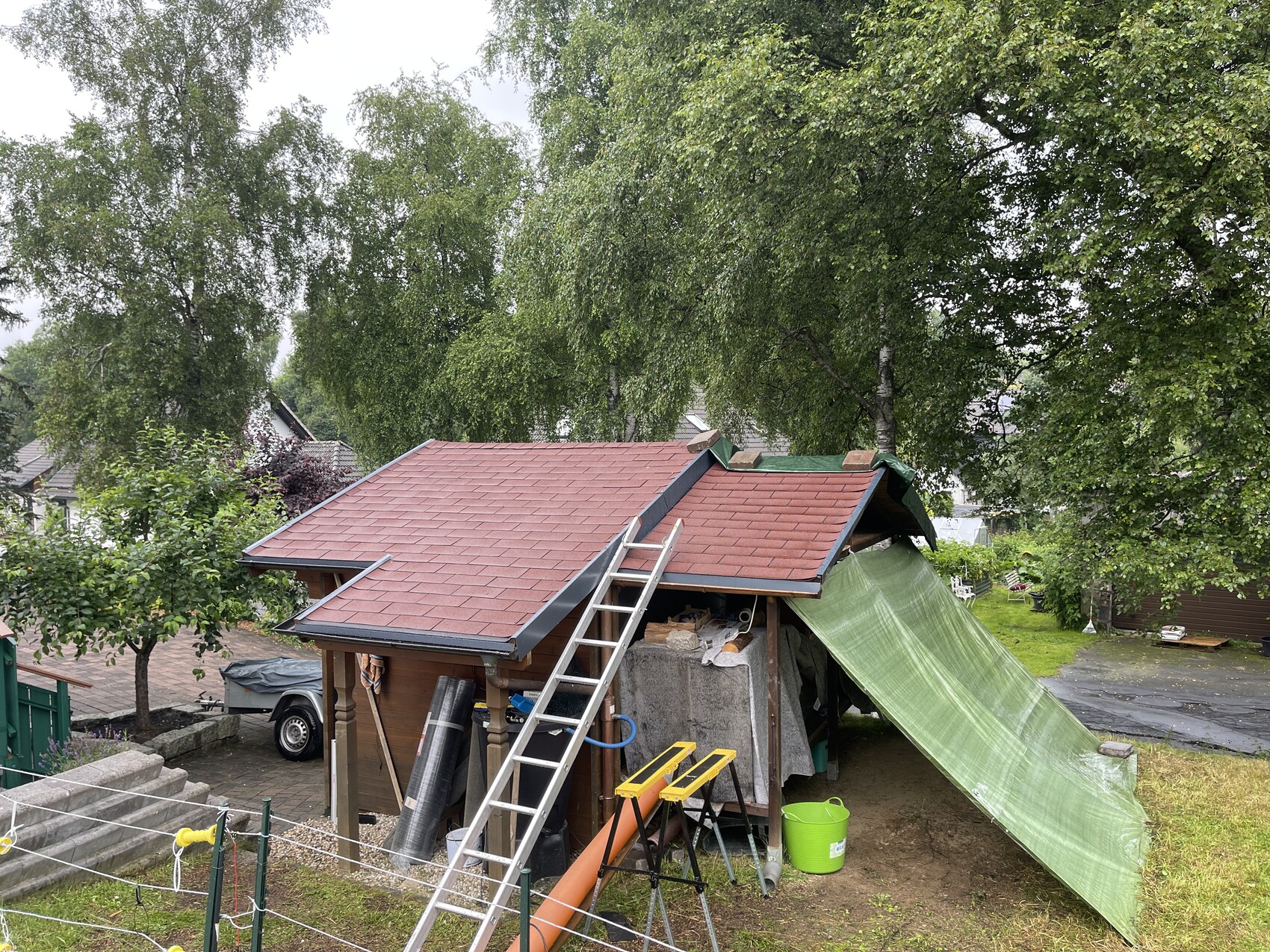 Kleines Holzhaus mit rotem Dach, Leiter davor, grüne Plane als Überdachung im Garten.