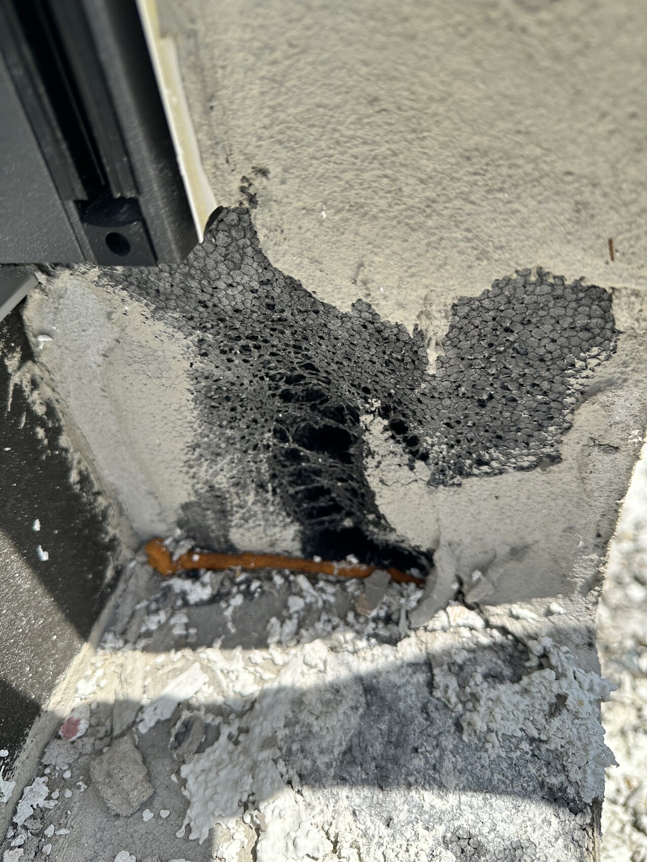 Rissige Betonwand mit offenporigem schwarzen Schaum, Kupferrohr liegt darunter.