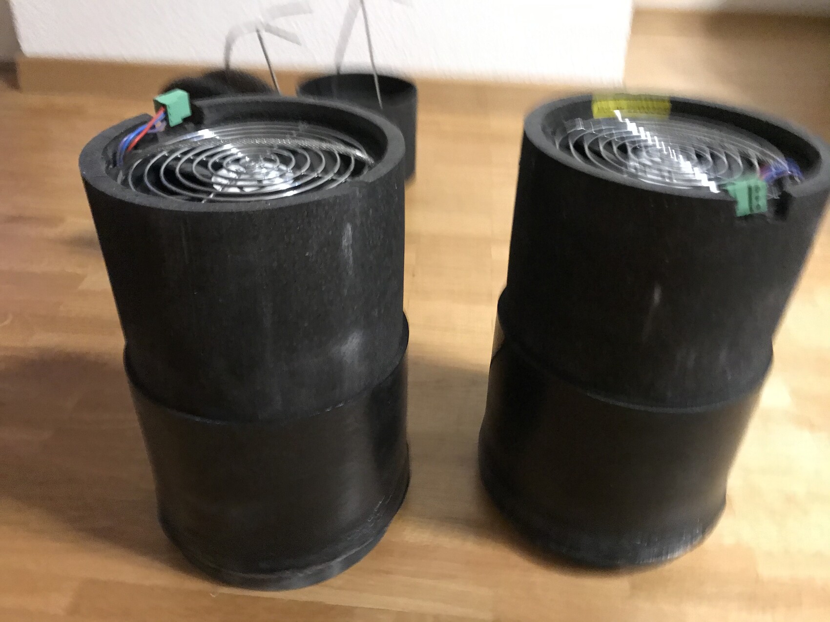 Zwei schwarze Zylinder mit spiralförmigem Metallgitter oben, grüne Stecker, auf Holztisch.