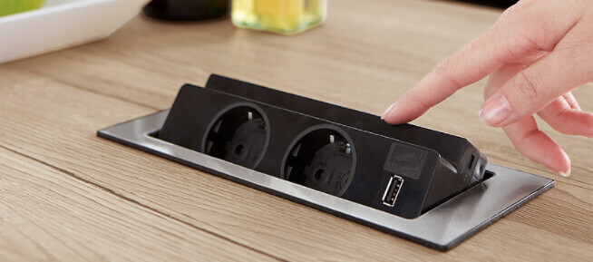 Schwarze Steckdosenleiste mit zwei runden EU-Steckdosen und USB-Port auf Holzplatte; Hand daneben