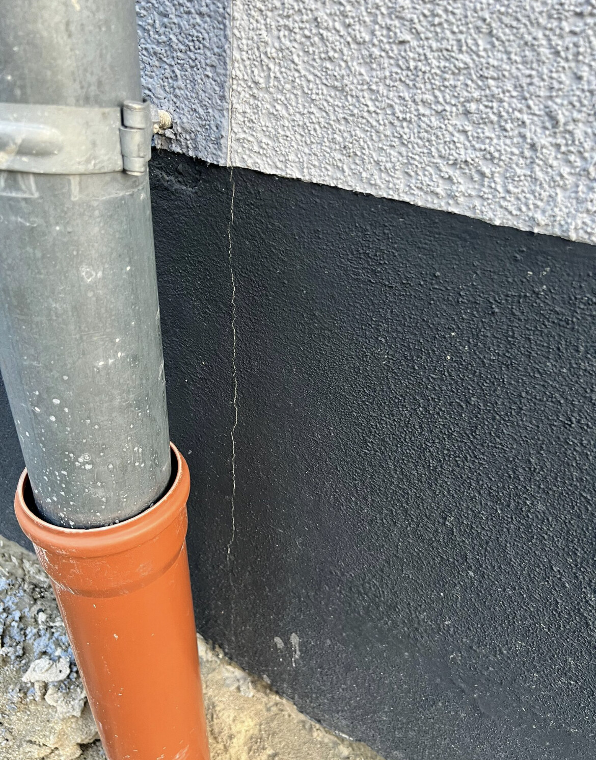 Außenansicht: graues Fallrohr an der Wand, schwarze Abdichtung unten; oranges PVC-Rohr am Boden.