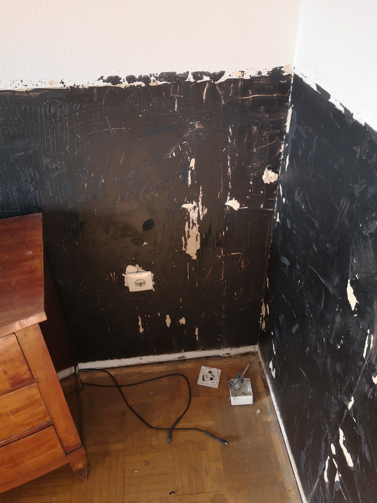 Eckszene: Schwarze Wand mit Abplatzern, Holzkommode links, Kabel auf dem Boden.