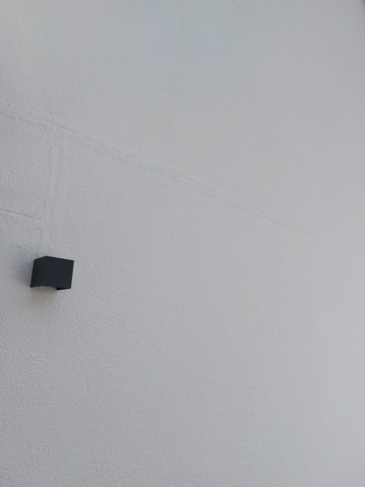 Weiße, grob-texturierte Wand mit einer kleinen schwarzen Kubus-Wandlampe links unten.