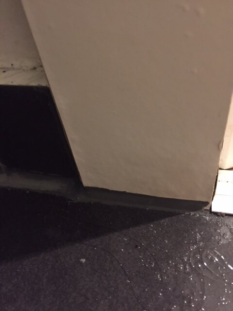 Ecke eines Raums mit schwarzem Bodenbelag, weißer Wand und Fußleiste.