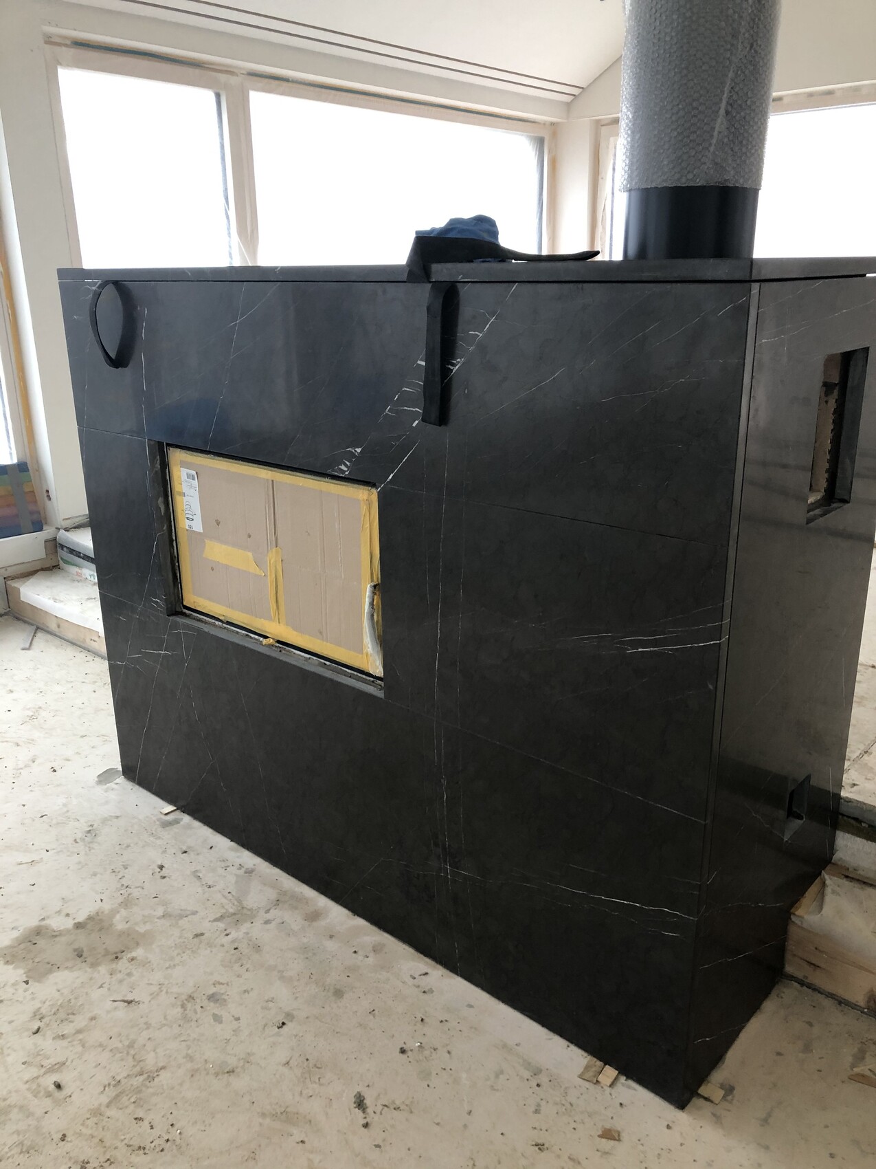 Großer schwarzer Kaminofen in Marmordesign mit Rohrführung oben; Kartonverpackung in Frontöffnung.