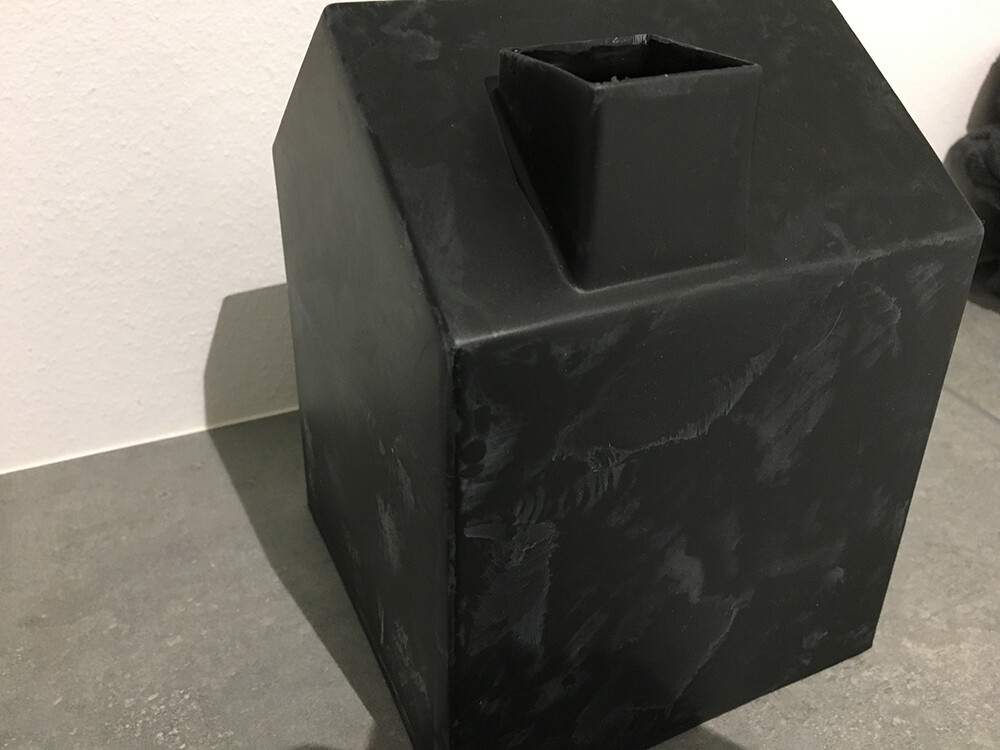 Schwarzer quadratischer Block mit hohem rechteckigem Auslass auf der Oberseite, auf grauem Boden.