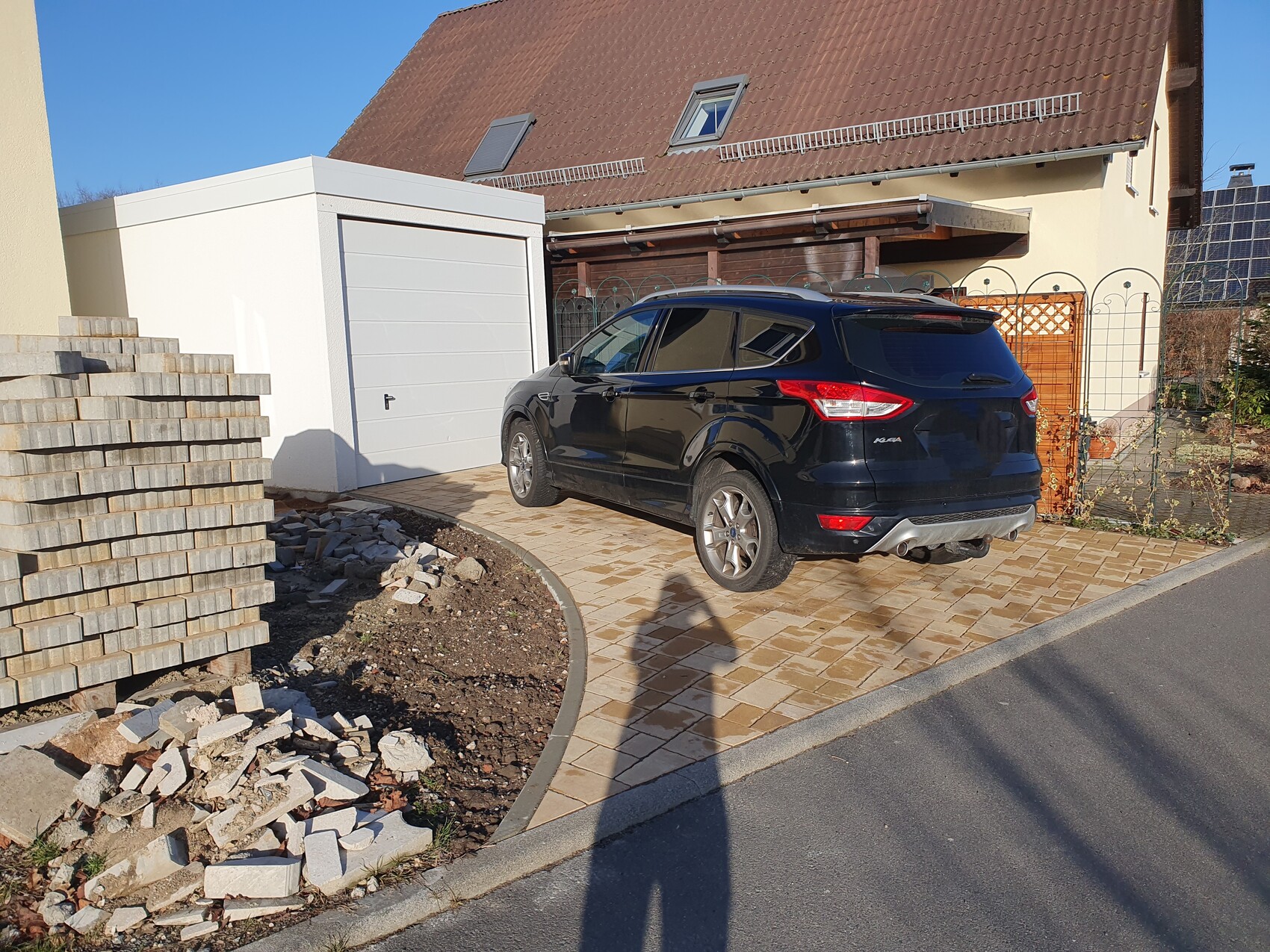 Schwarzer SUV in gepflasterter Einfahrt vor weißer Garage; Baureste links.