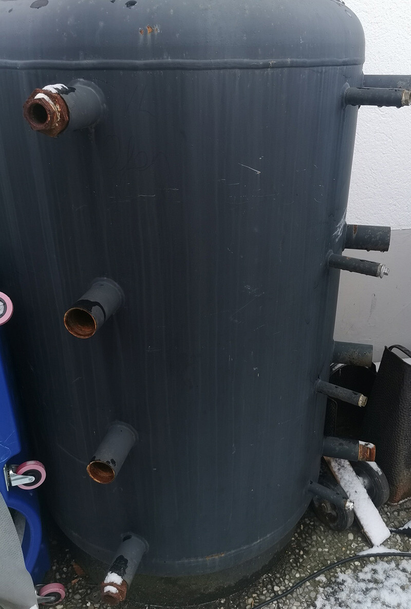 Großer dunkler Zylinderbehälter im Freien mit mehreren verrosteten Rohranschlüssen seitlich.