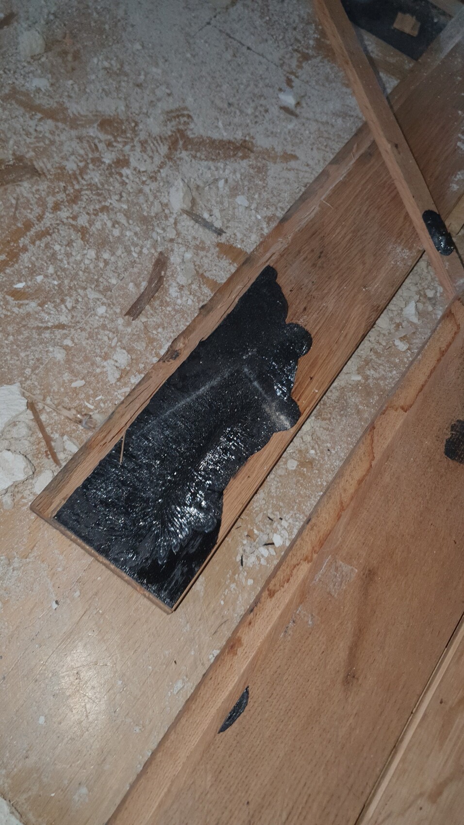 Schwarze Bitumenabdichtung auf einer Holzlatte in einer Baustellenszene mit Staub und Holzresten.