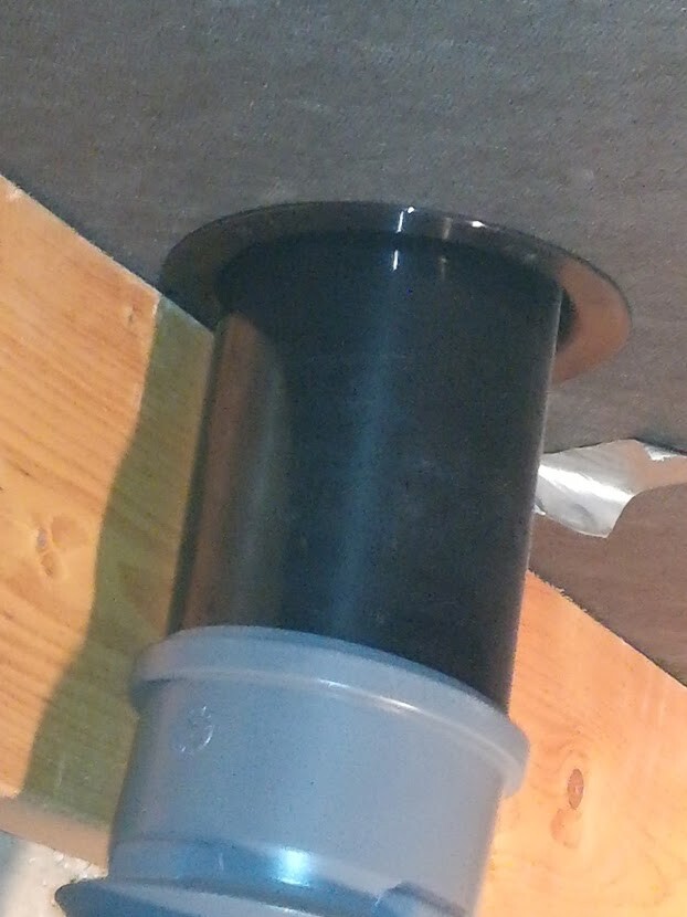 Schwarzer Rohrstutzen mit Flansch-Oberteil und grauer PVC-Verbindung vor Holzwand.