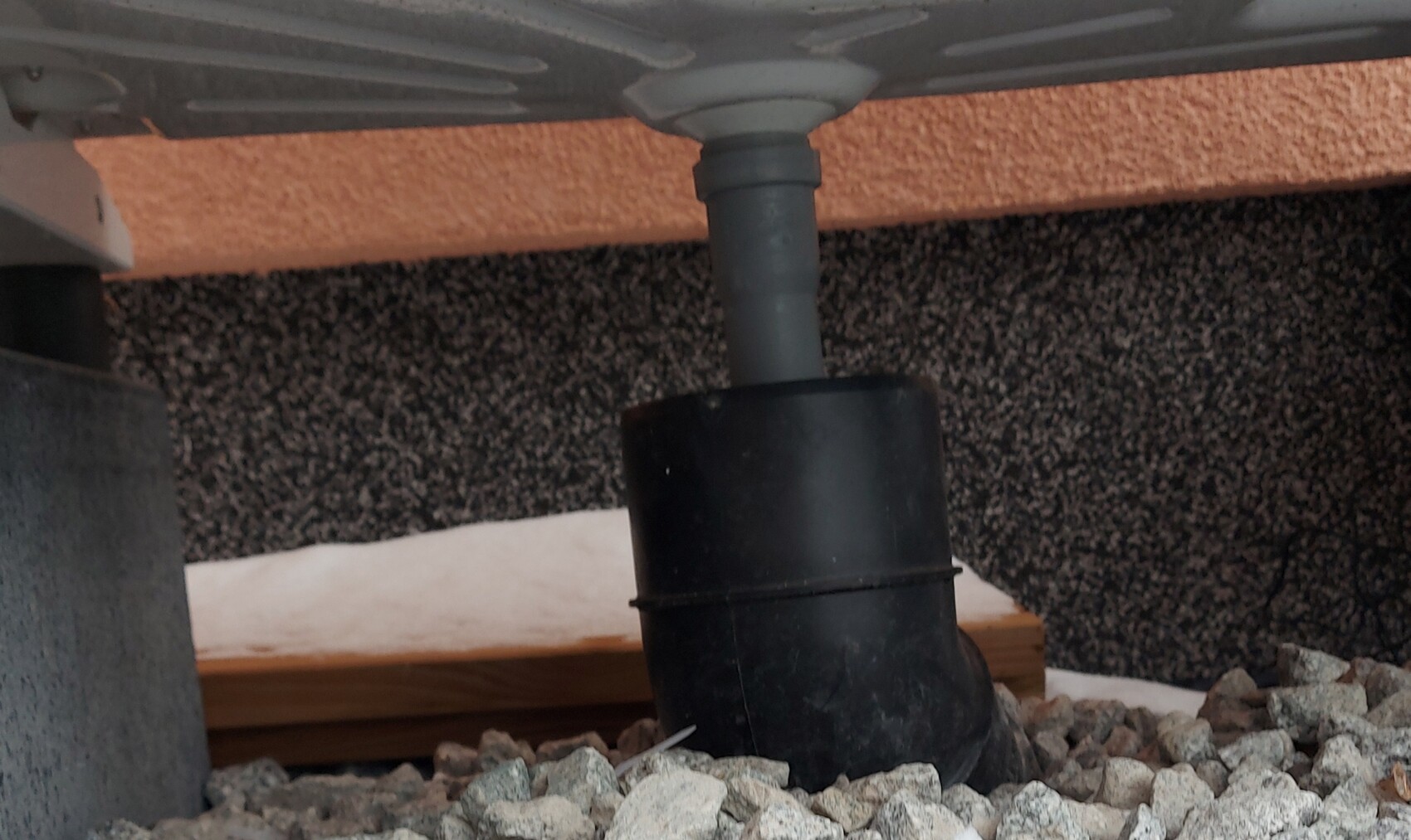Unteres Abflussrohr mit schwarzem Siphon, außerhalb eines Gebäudes, Kiesboden.