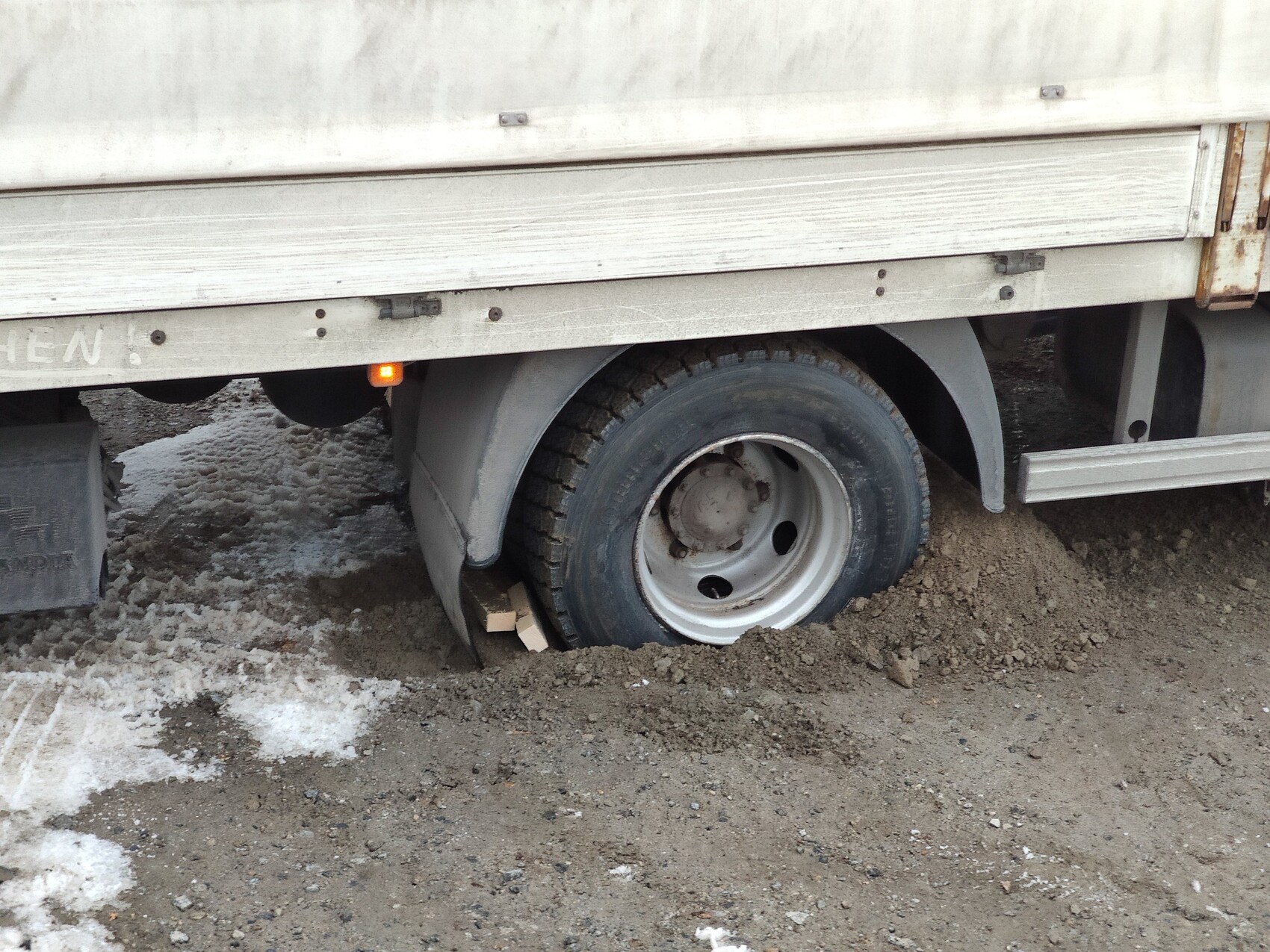 LKW-Anhänger mit Rad im Schlamm, Holzklotz unten, staubige Baustellenumgebung.