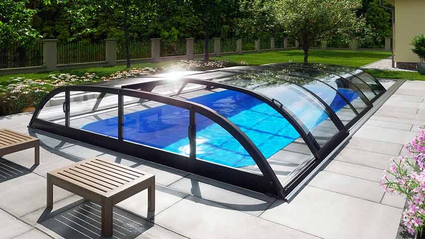 Kuppelförmige Glas-Poolabdeckung über blauem Becken auf Terrasse mit Garten und Bänken.