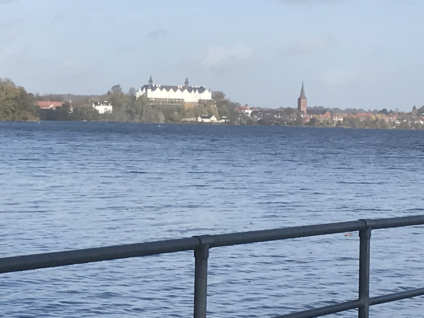 Blick über einen See auf eine Stadt mit Kirchturm und weißen Gebäuden; Geländer im Vordergrund.