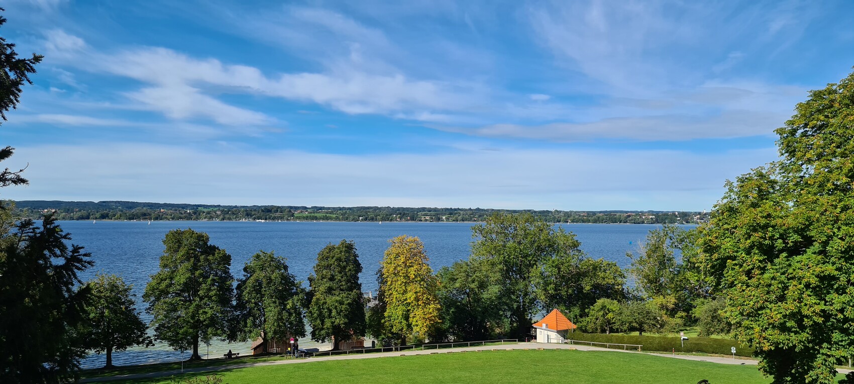 Parkblick auf einen See: grüne Wiese, Bäume am Ufer, kleine Hütte mit orangefarbenem Dach