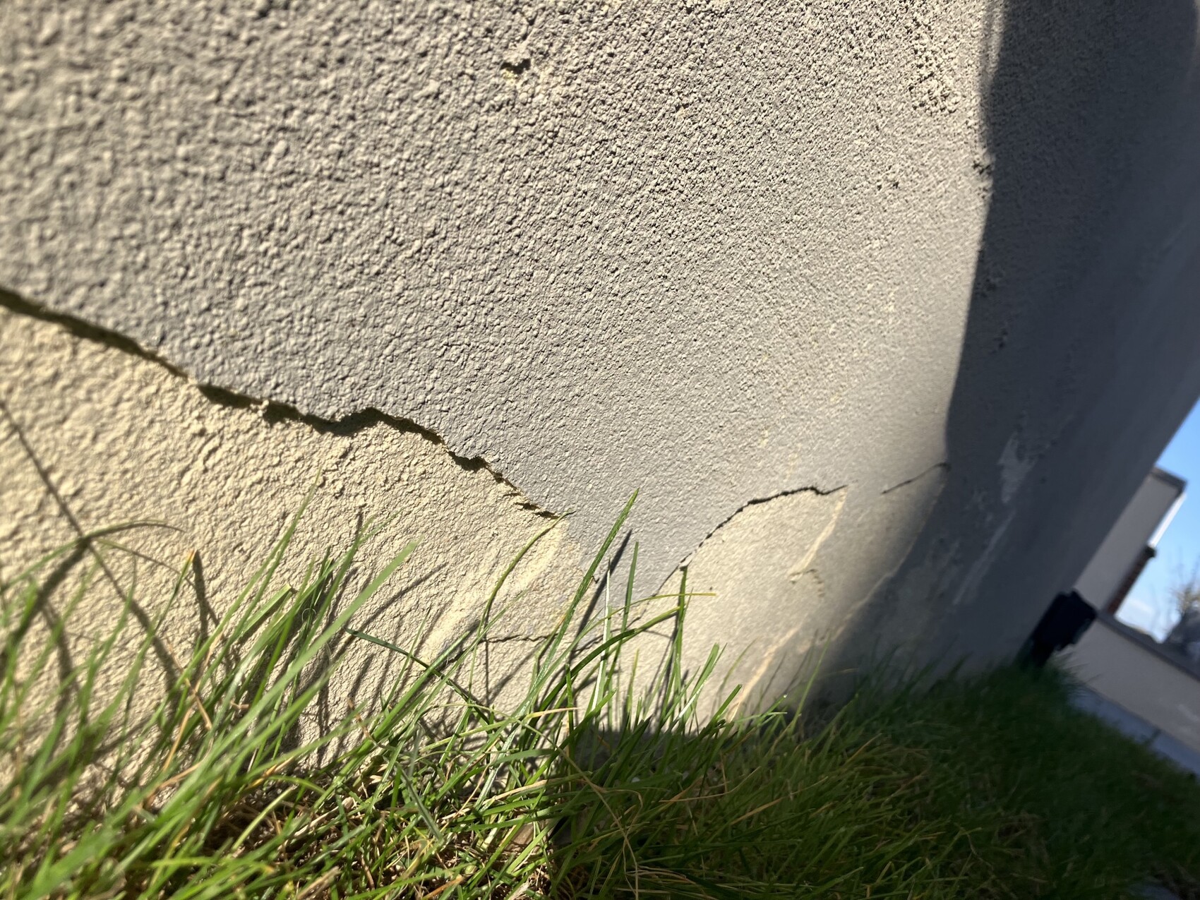 Rissige Betonwand mit Gras am Fuß; Gebäudekante im Hintergrund.