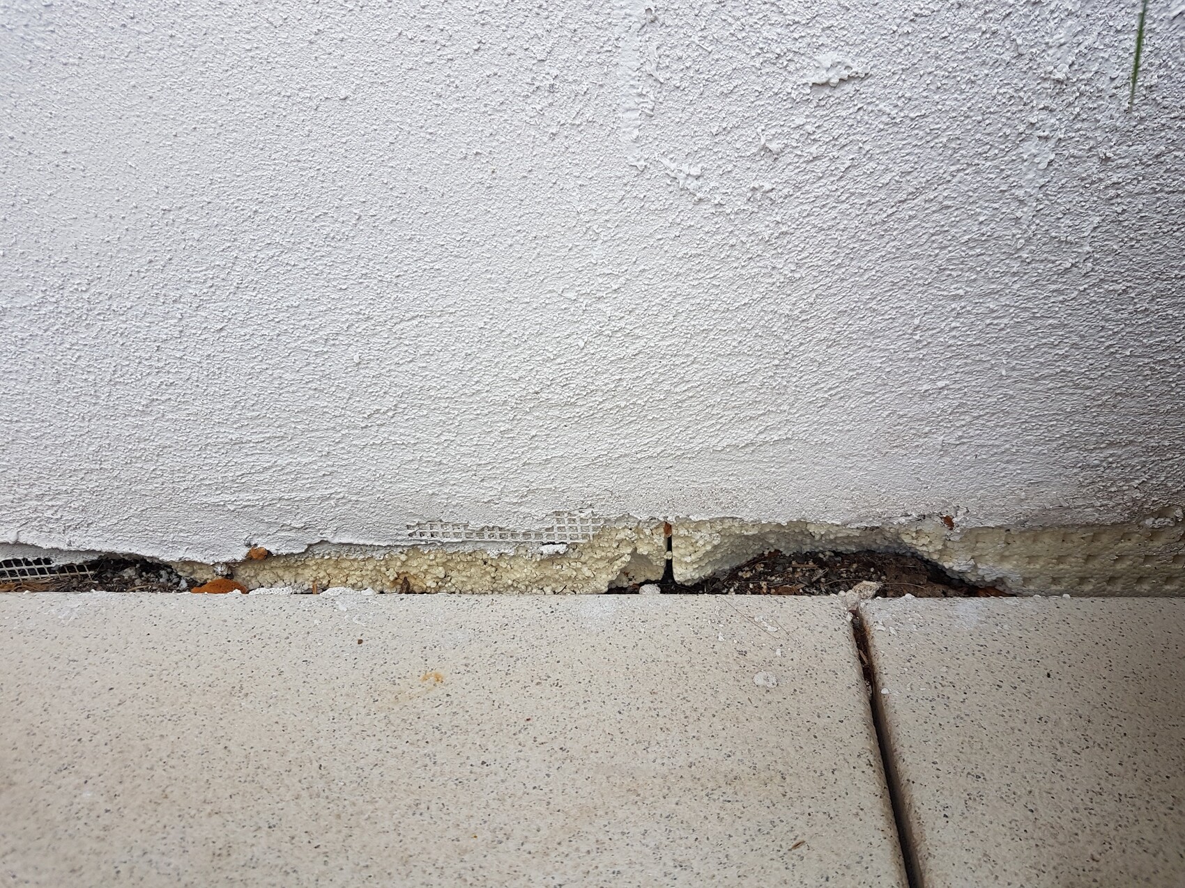 Weiße verputzte Wand mit Bodenriss und sichtbarer Dämmung zwischen Wand und Fliesen.