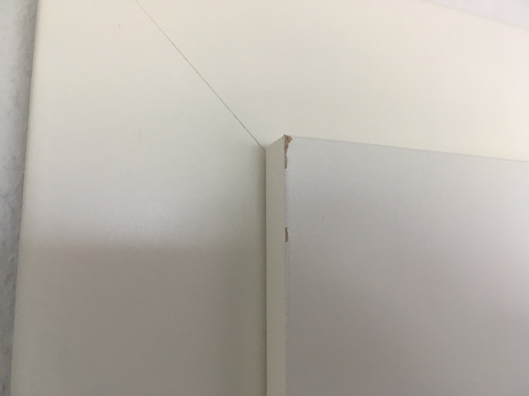Nahaufnahme einer Innenwand-Ecke: weiße Wand neben hellgrauem Paneel; kleine Absplitterung.