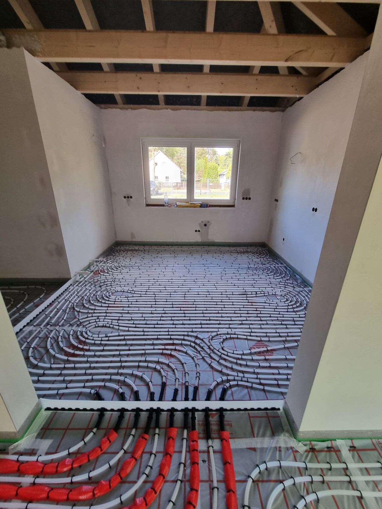 Baustellenraum mit Fußbodenheizung: rote Rohre in Schlaufen auf dem Boden, Fenster sichtbar.