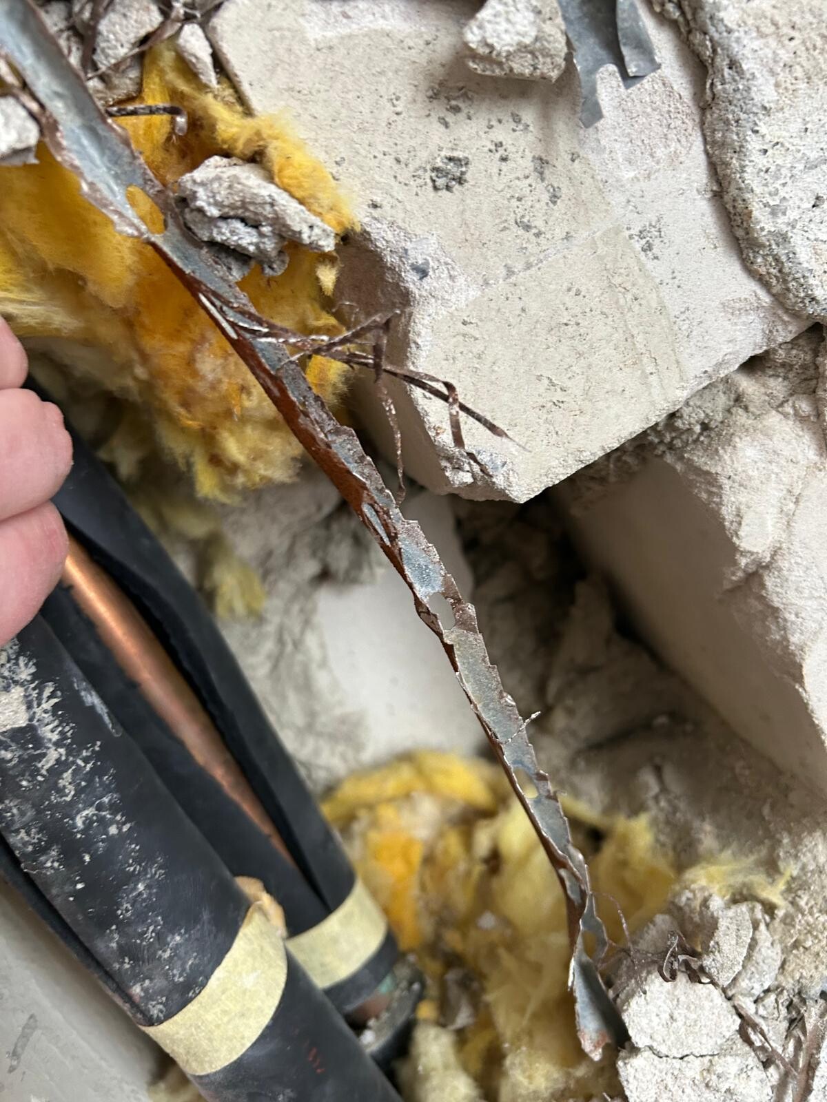 Aufgerissene Wand mit gelber Dämmung, rostigem Metallstreifen und Rohren.