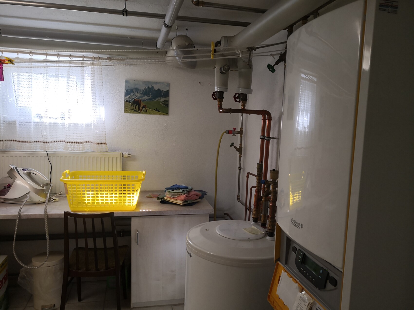 Waschküche mit Boiler, Kupferleitungen, Waschmaschine, gelbem Wäschekorb und Wäschestapel.