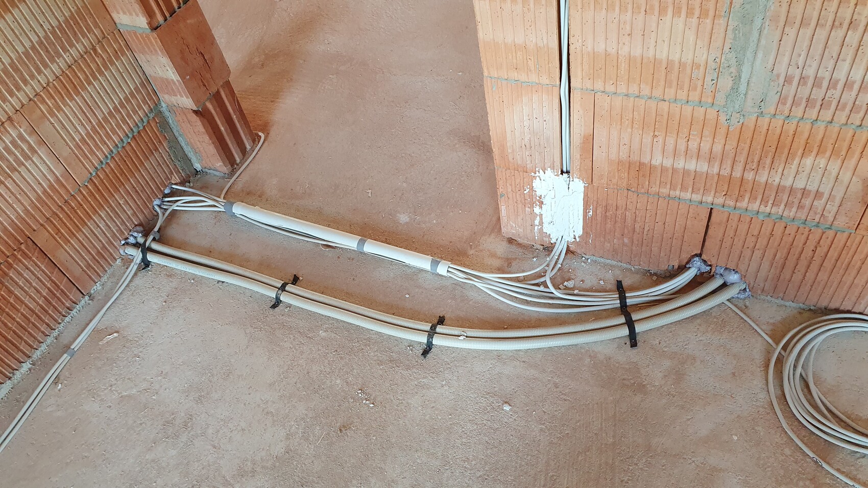Auf einer Baustelle verlaufen weiße Kabelrohre und Schläuche am Boden neben einer rohen Ziegelwand.