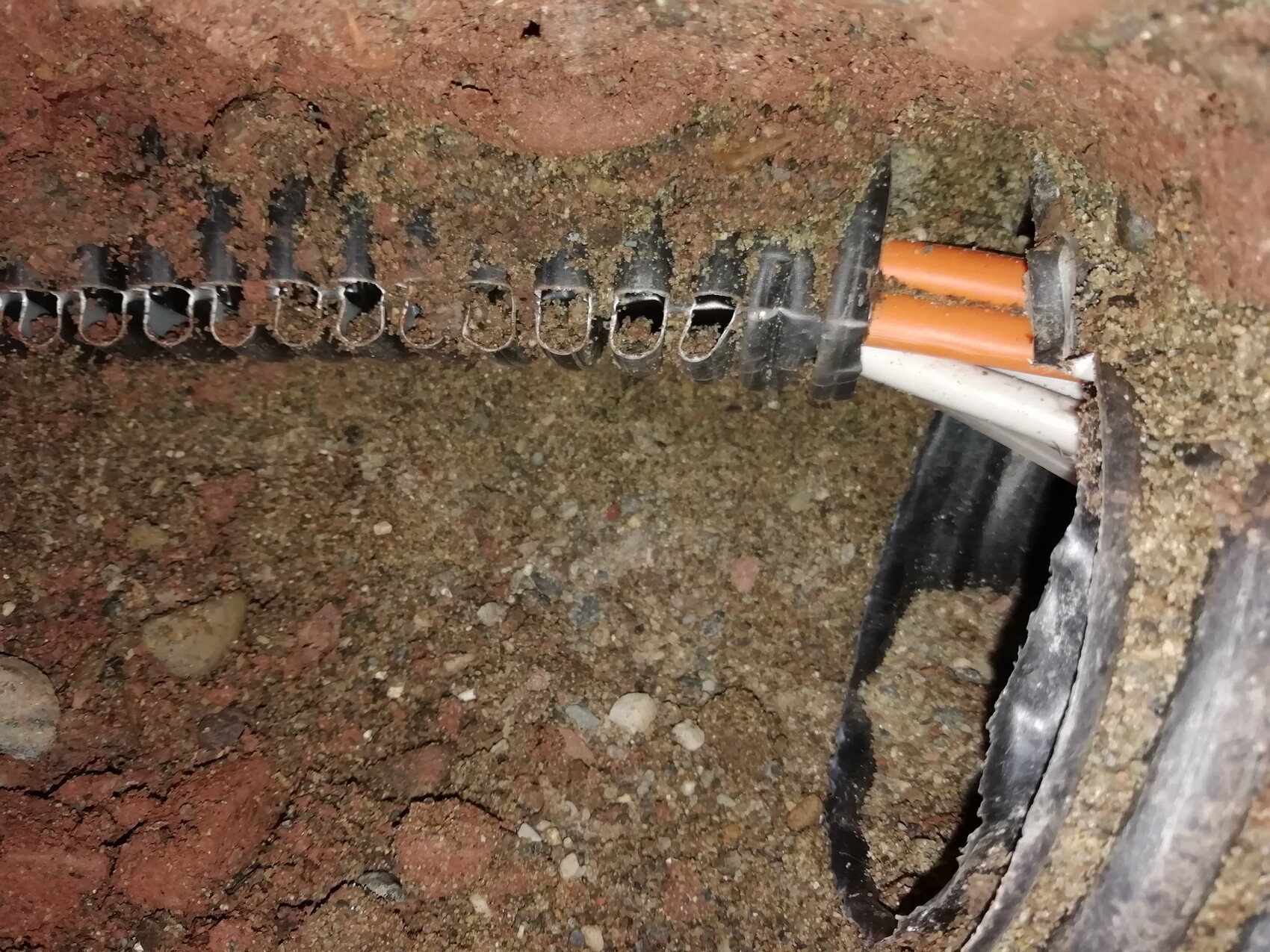 Offene Erdöffnung zeigt orange und weißes Kabel in beschädigter Leitung, umgeben von Staub.