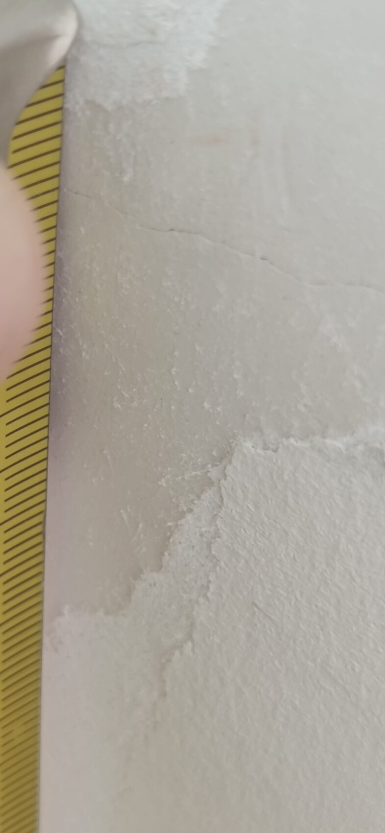 Nahaufnahme einer weißen Putzwand mit Rissen; links ein Maßband.