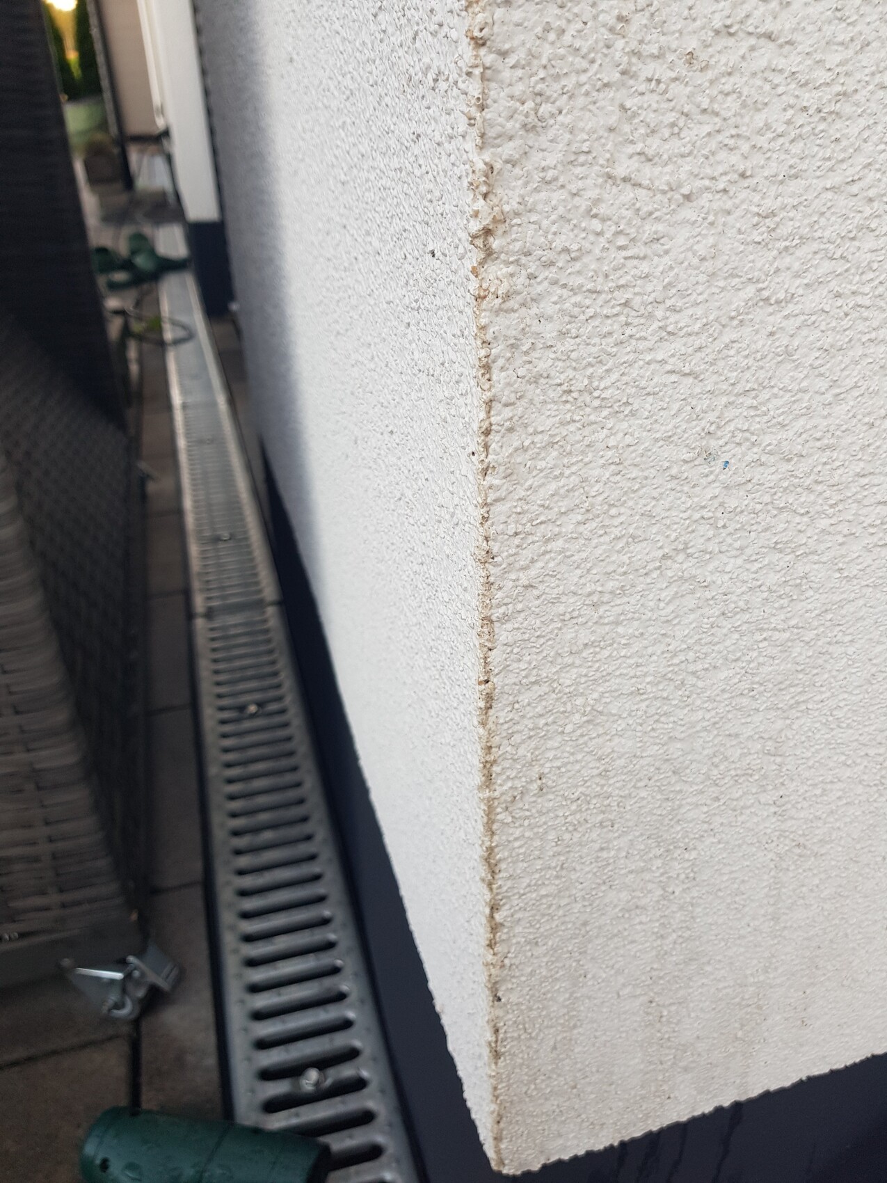 Weiß verputzte Außenwand mit vertikalem Riss am Eckbereich neben einer Metallrinne am Boden.
