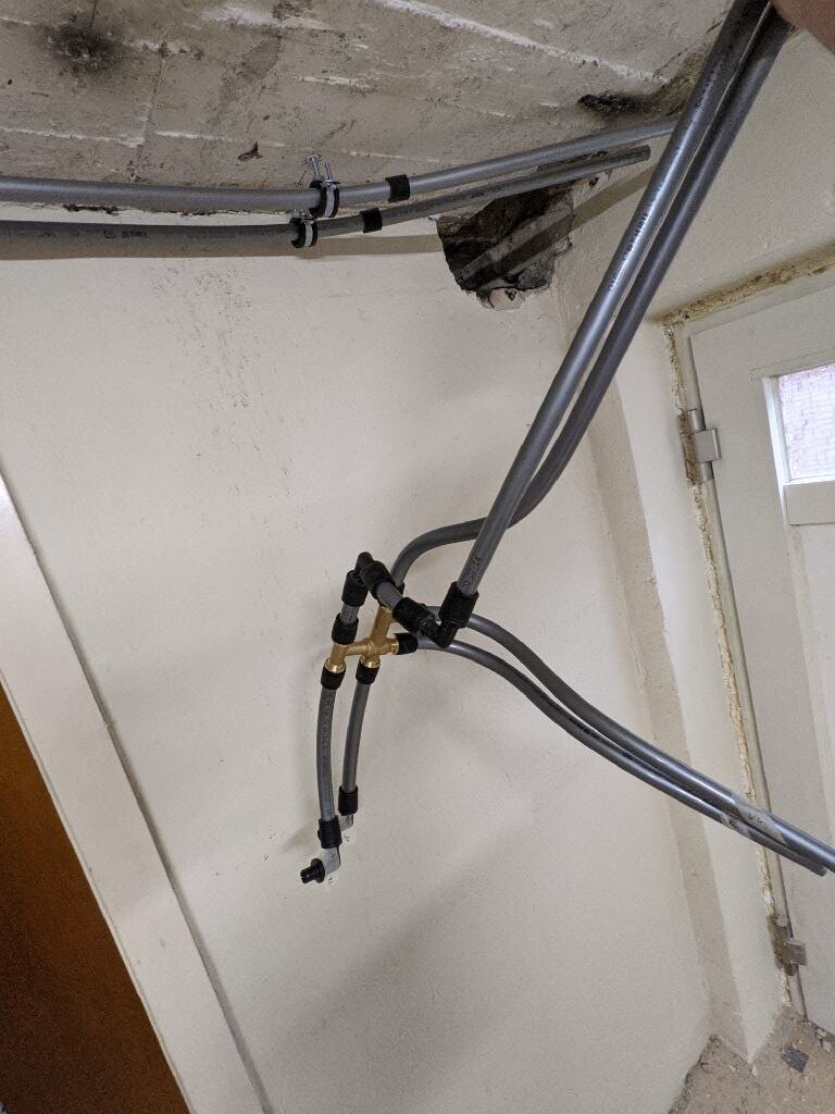Graue Leitungen und Schläuche hängen an der Wand, Messing-Verzweigstück, Loch in der Decke.