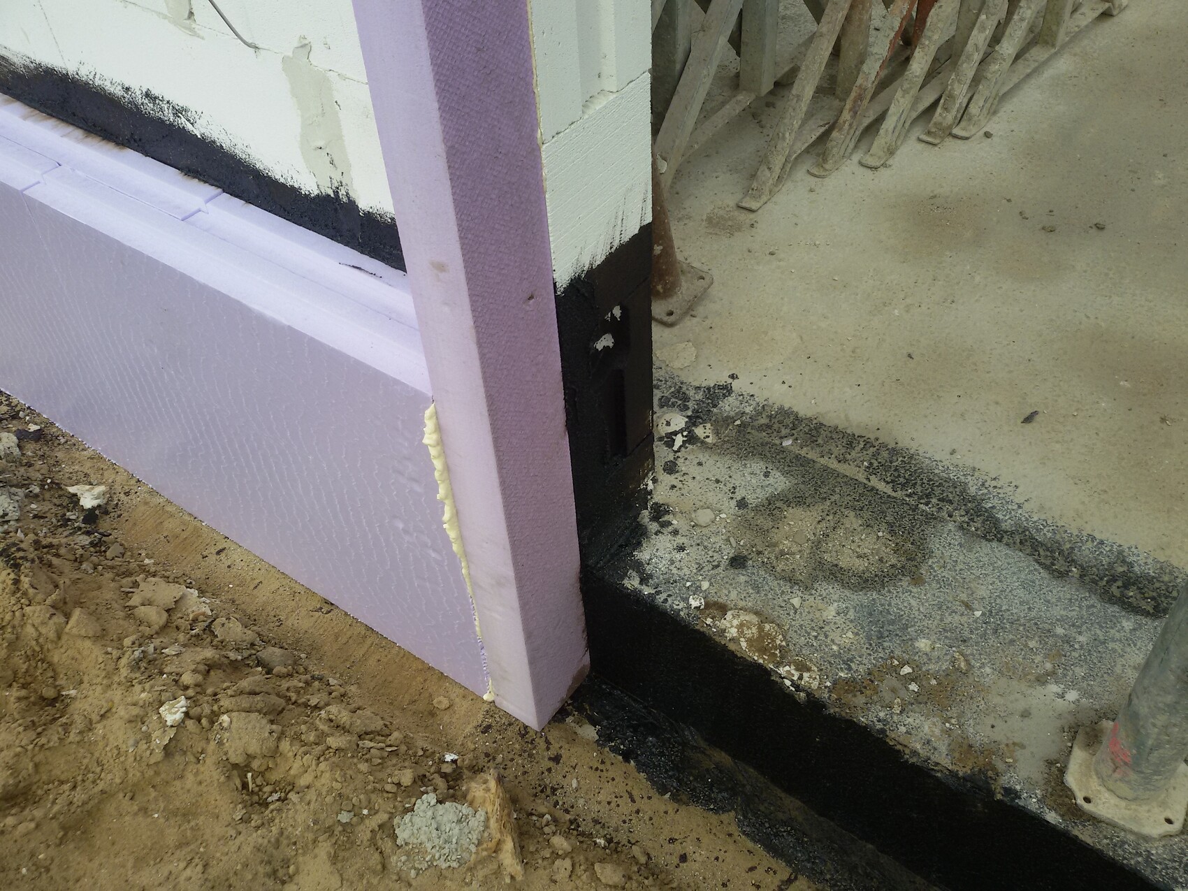 Außenwand-Baustelle mit lila Dämmplatten, schwarze Fundamentabdichtung, Boden aus Beton und Erde.
