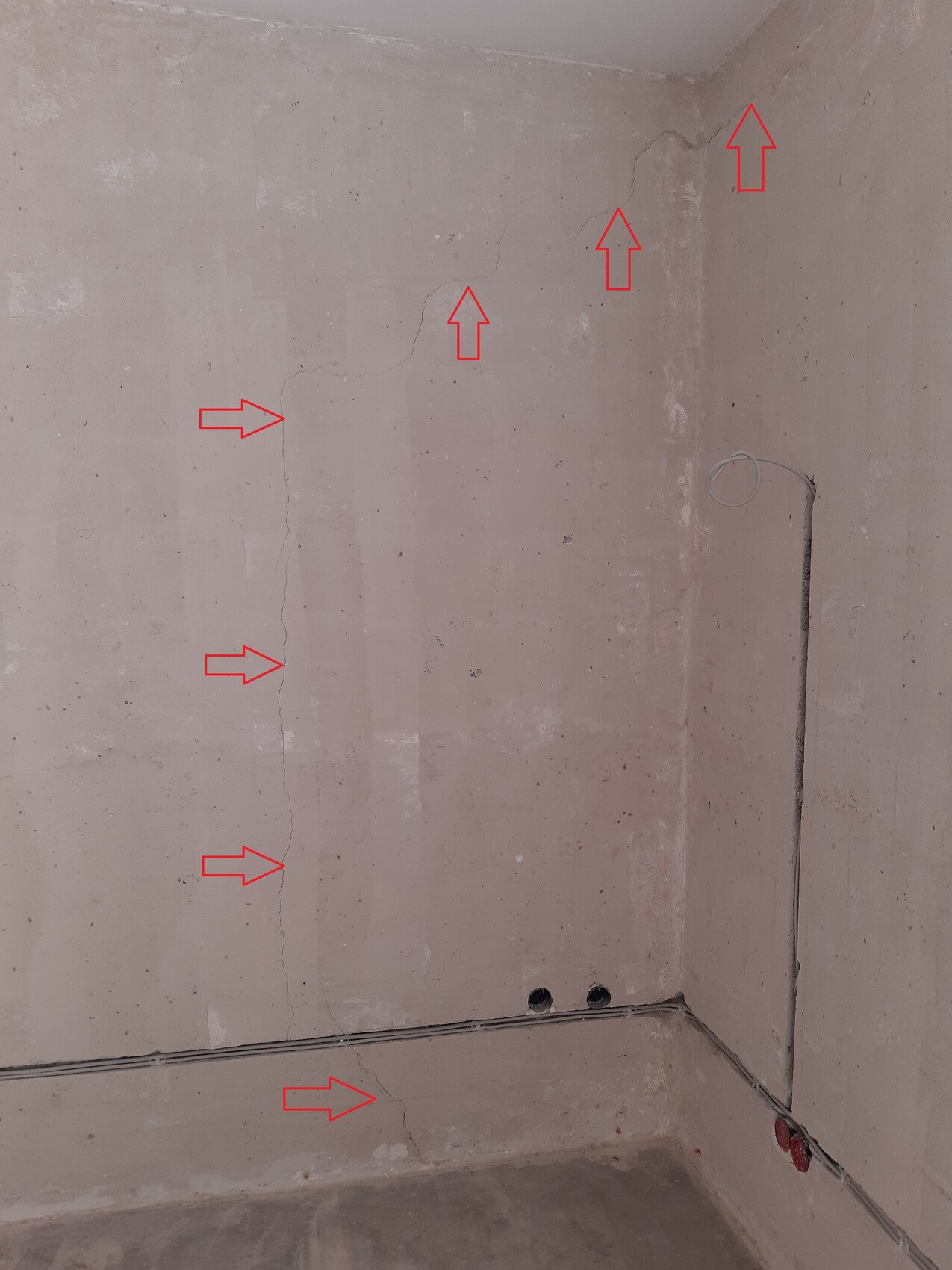 Mehrere Risse in roher Betonwand; rote Pfeile markieren Risse, Kabeldurchführung und Bodenlöcher.
