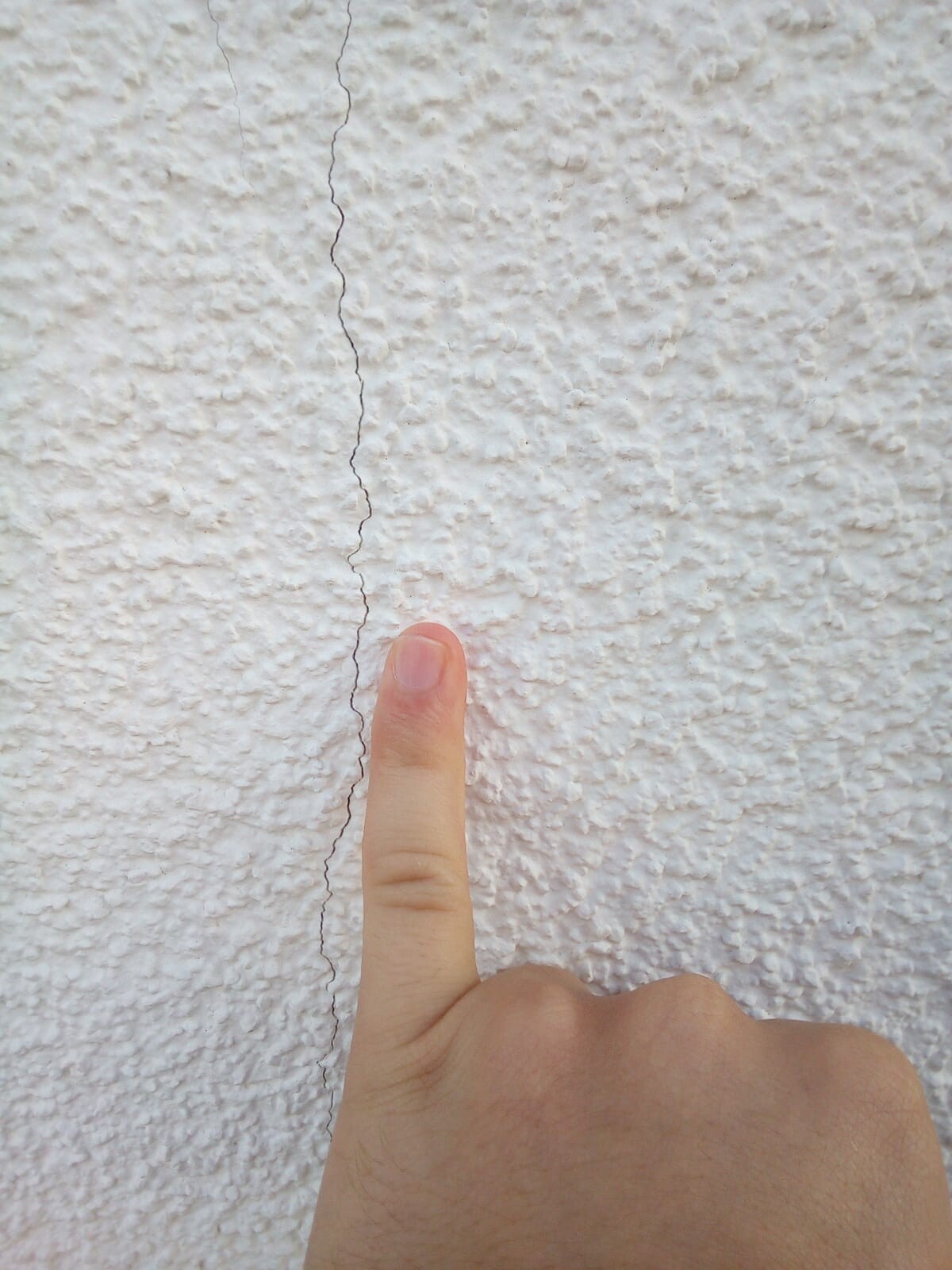 Finger zeigt auf feinen Riss in weißer, rauer Wand (Nahaufnahme).