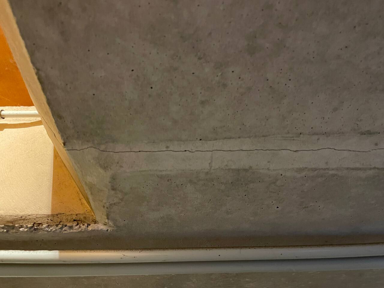 Riss in grauer Betonwand; linke orange Ecke, unten weiße Rohrleitung sichtbar.