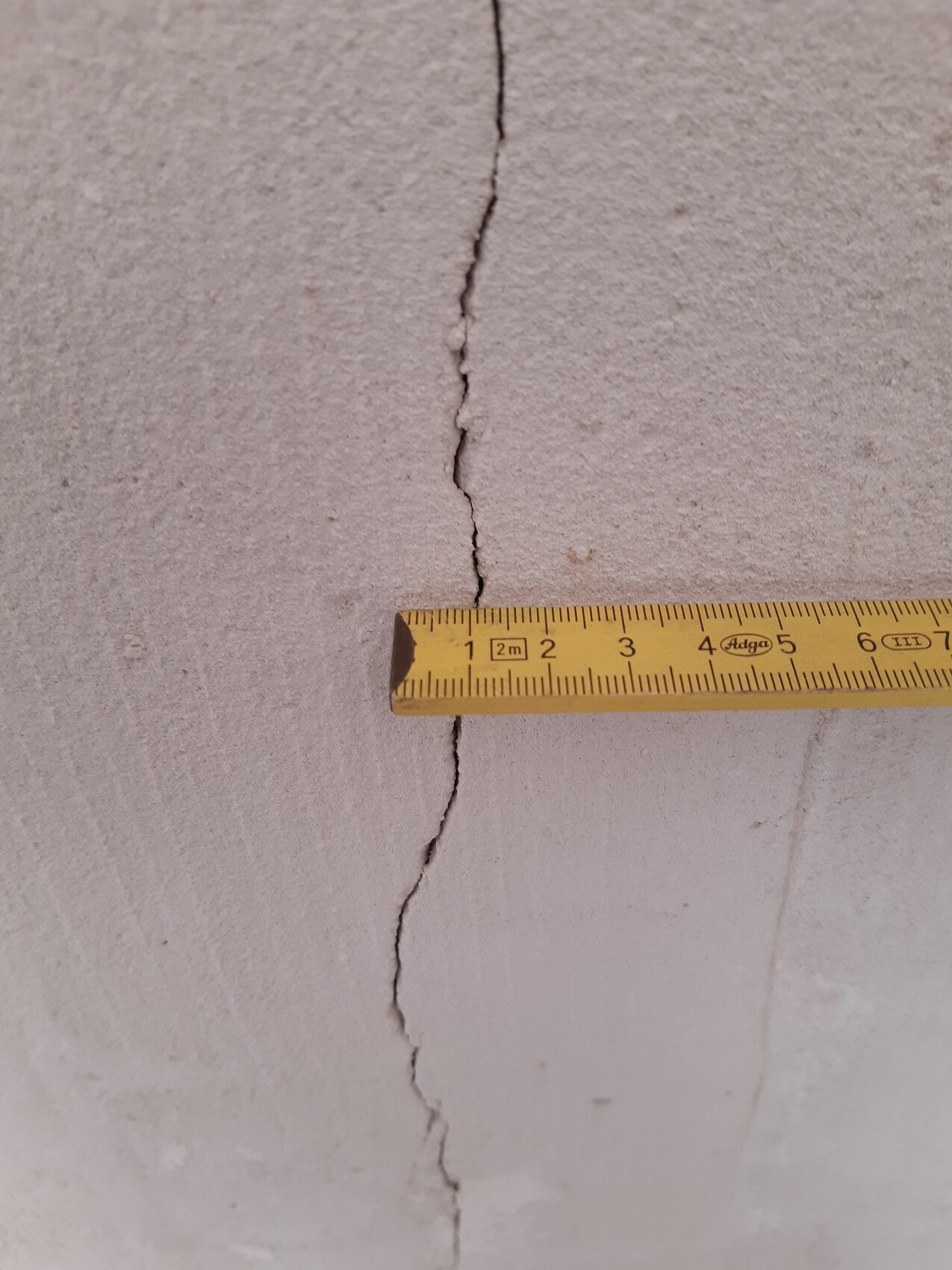 Riss im grauen Beton, daneben liegt ein gelbes Maßband, das die Spalte misst.