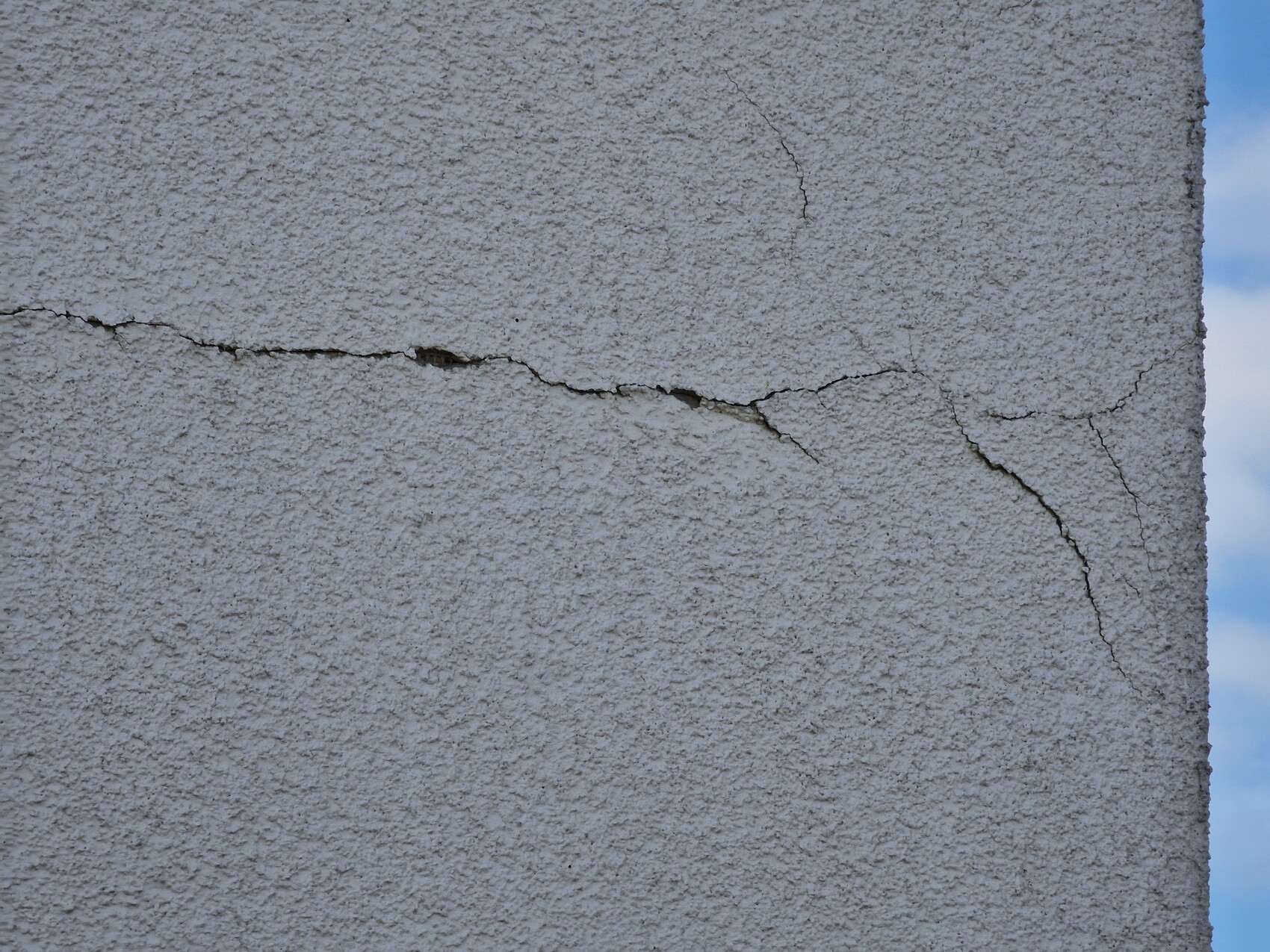 Nahaufnahme einer rauen, verputzten Wand mit Rissen; blauer Himmel am rechten Rand.