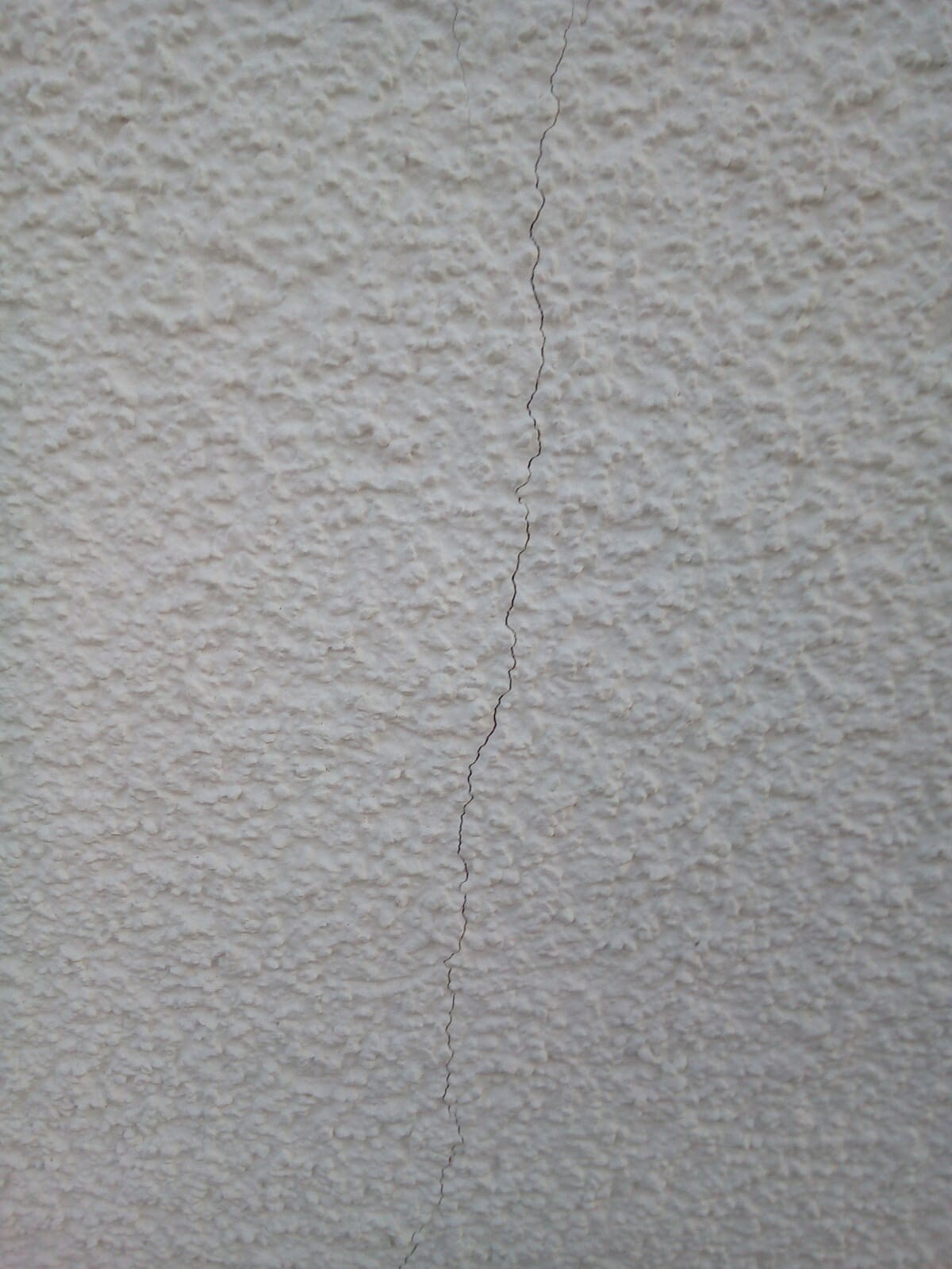 Riss in einer rauen weißen Wand, zentral von oben nach unten verlaufend.