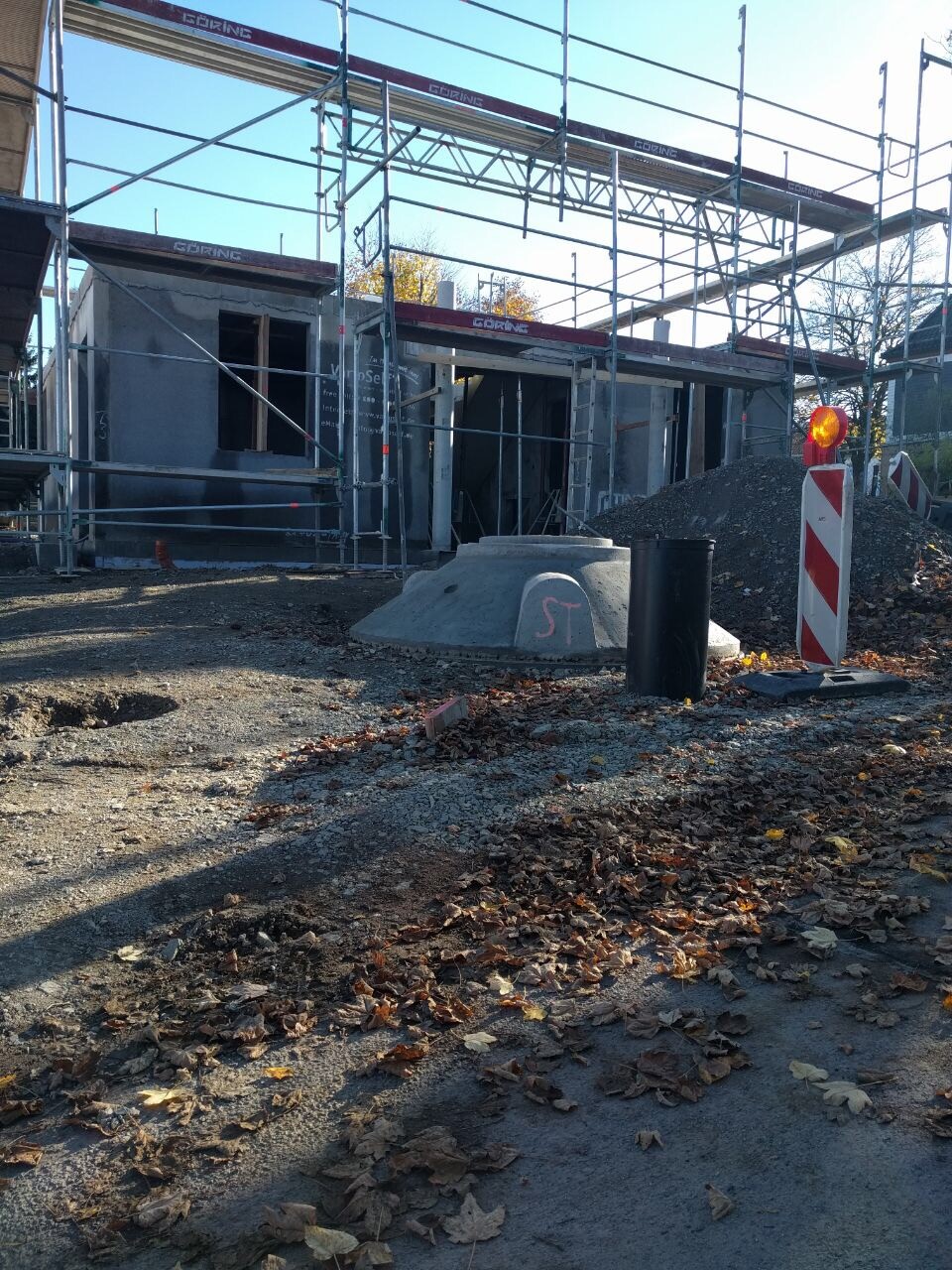 Baustelle mit Gerüst um unfertiges Gebäude; Kieshaufen, runder Betondeckel, Absperrpfosten.