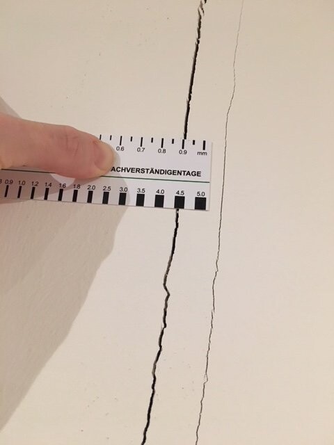 Nahaufnahme einer hellen Wand mit Risslinie; Hand hält Zentimeterlineal am Riss.