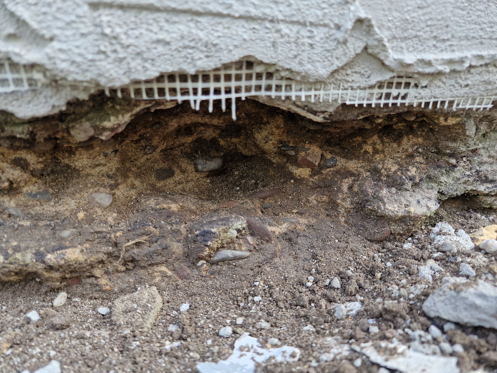 Unter einer beschädigten Wand liegt erdiger Boden mit Kieseln; Putz bricht ab.