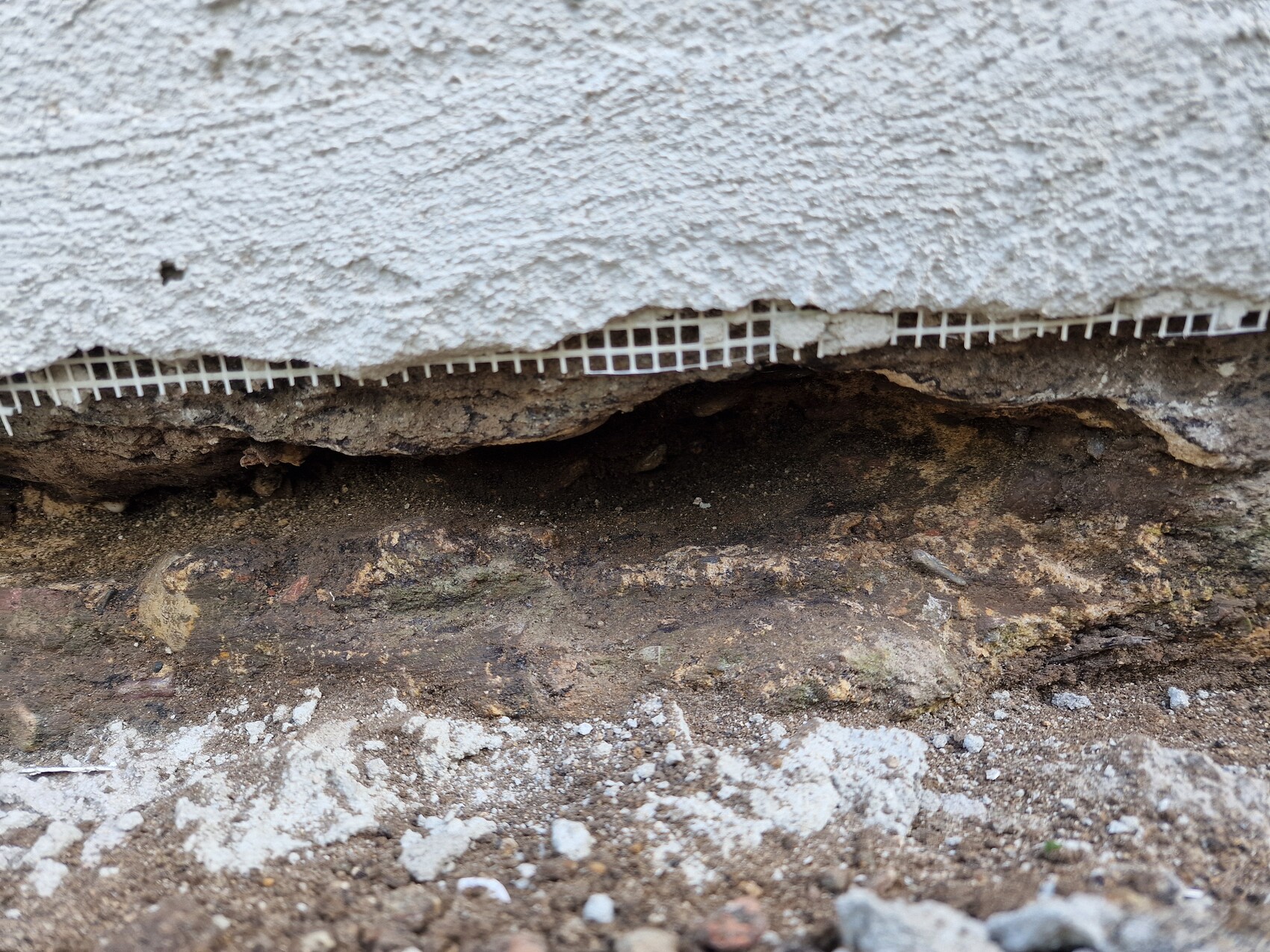 Unterseite einer verputzten Wand; freigelegtes Erdreich und bröckelnder Putz am Fundament.