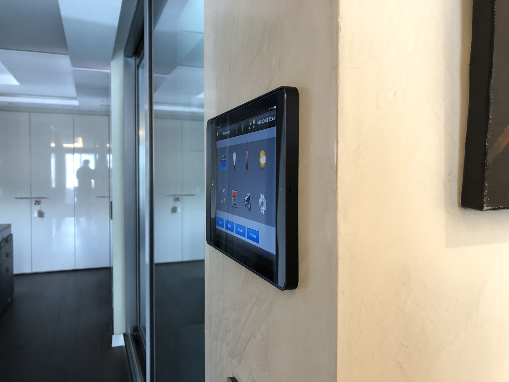 Auf einer beige Wand montiertes Tablet mit App-Icons; Glasflur im Hintergrund, Silhouette einer Person.