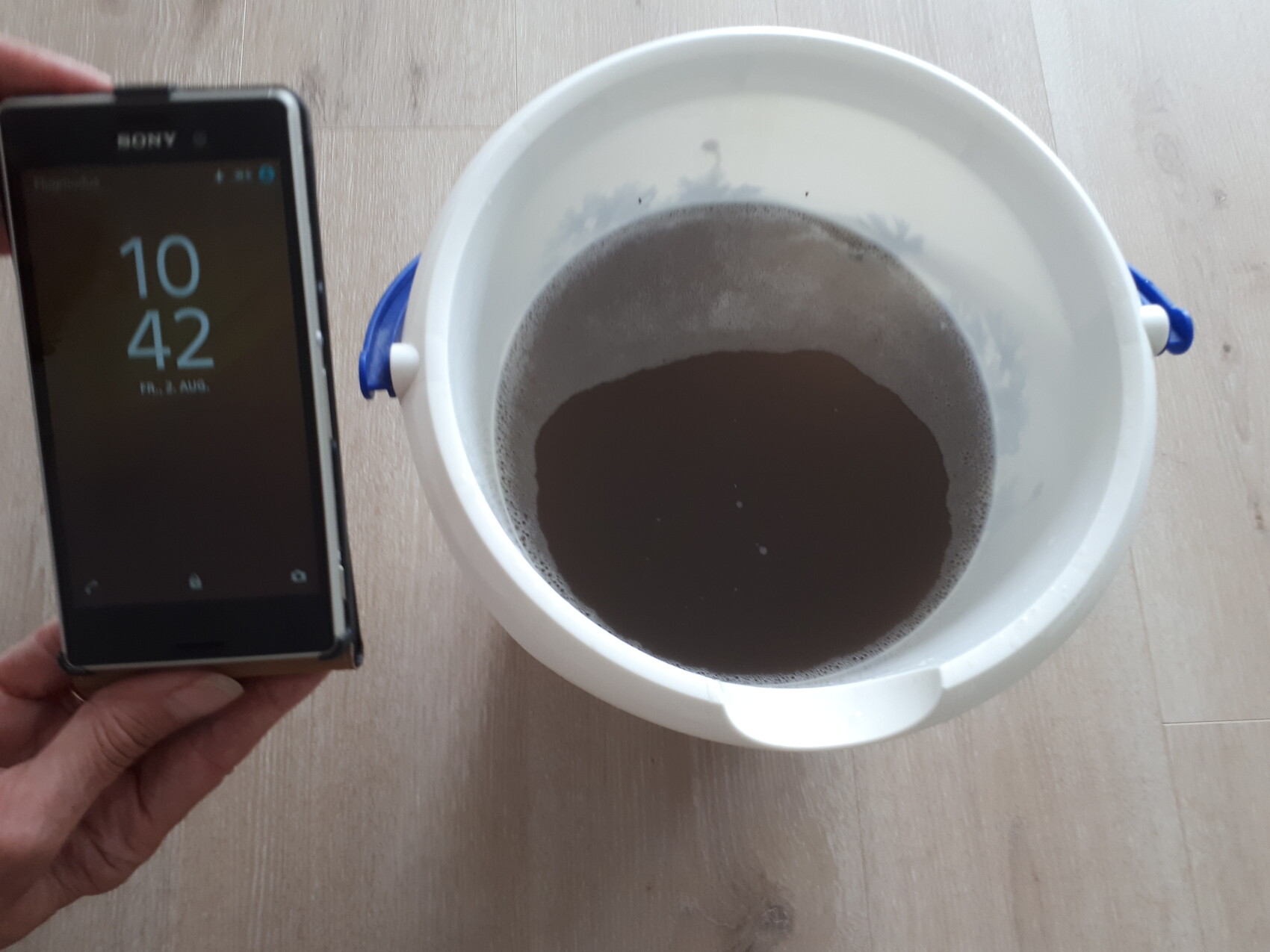 Person hält Smartphone neben weißem Eimer mit dunkler Flüssigkeit; Smartphone zeigt 10:42.