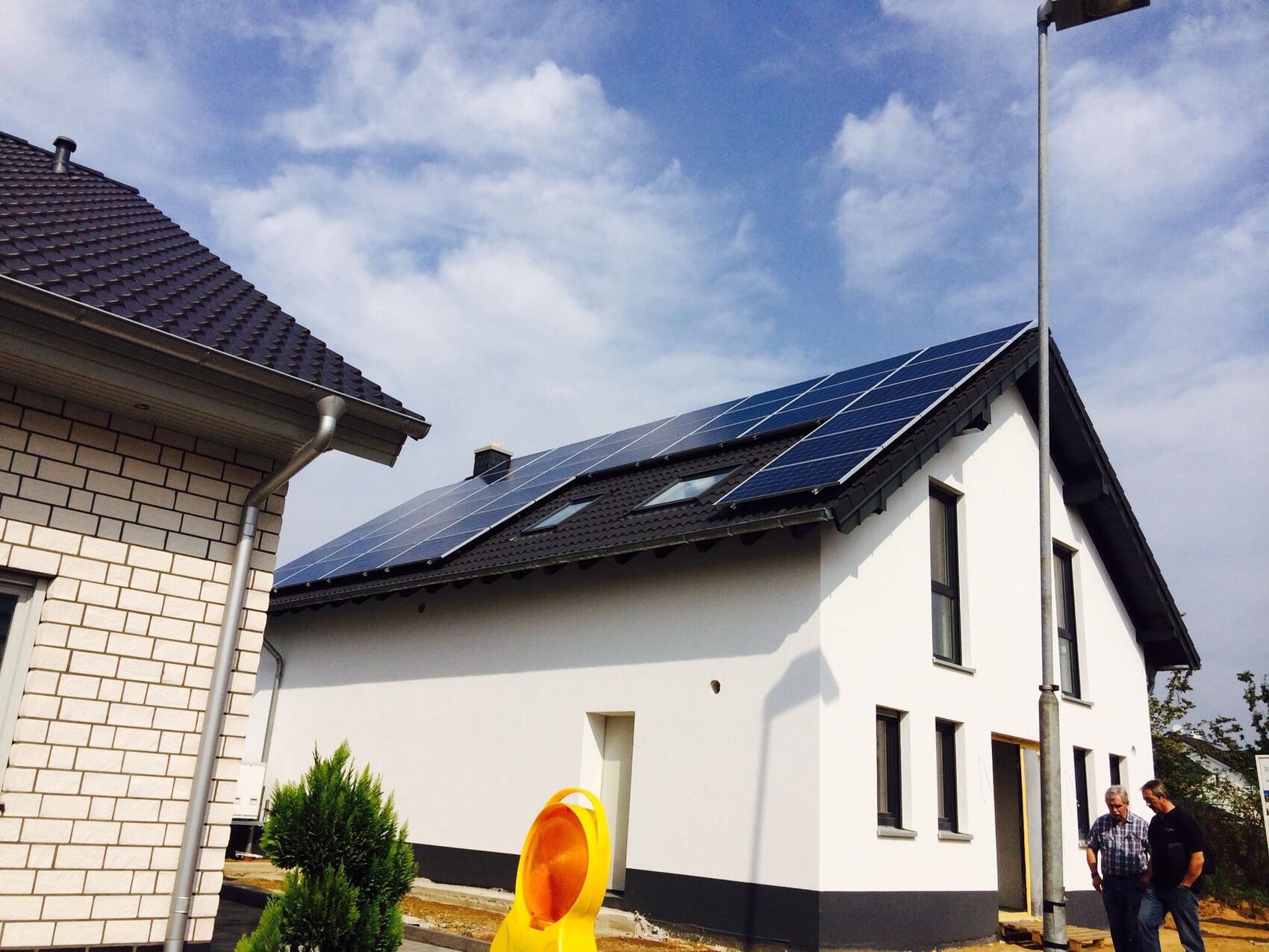 Modernes Wohnhaus mit Solarzellen-Dach; zwei Männer vor dem Eingang, Gelber Kegel im Vordergrund.