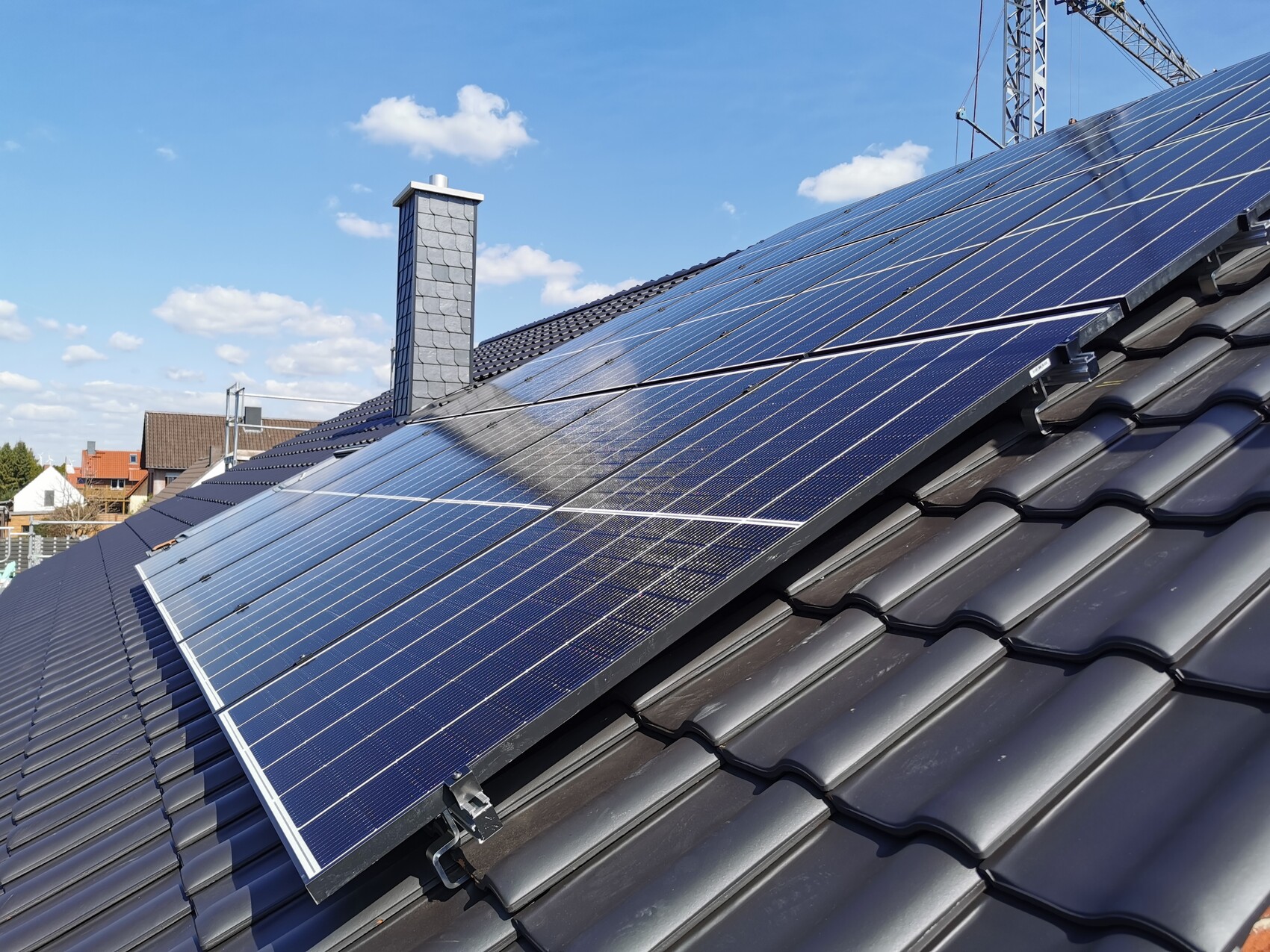 Dach mit Solarmodulen auf dunkelgrauen Ziegeln, Kamin und Baukran am blauen Himmel.