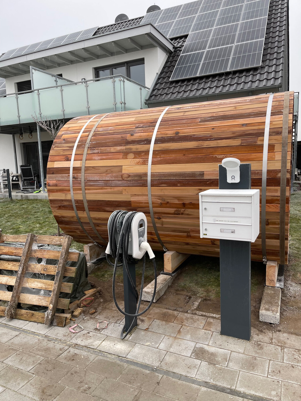 Großes Holzfass-Hot-Tub im Garten vor modernem Haus mit Solarzellen auf dem Dach.