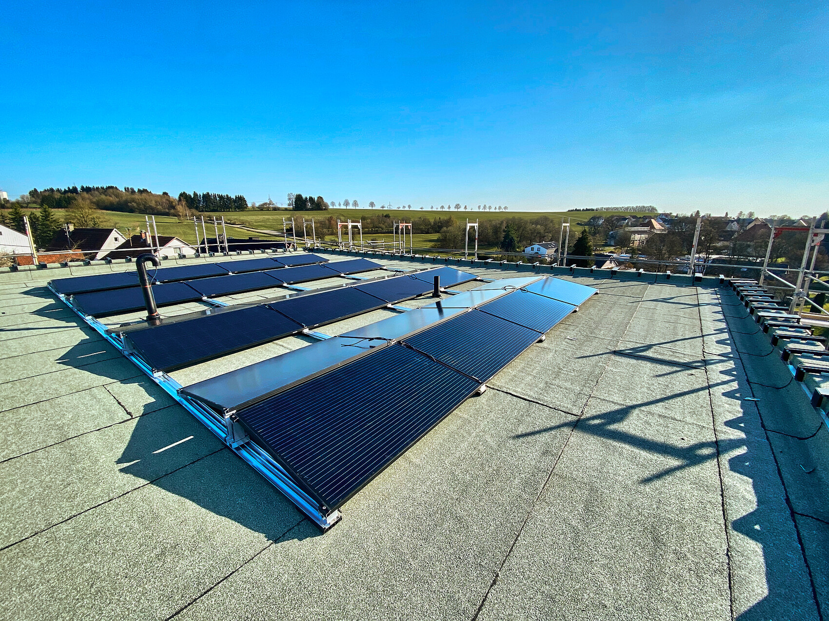 Flachdach mit Solarmodulen, Reihen von Panels; ländliche Häuser und Felder im Hintergrund.