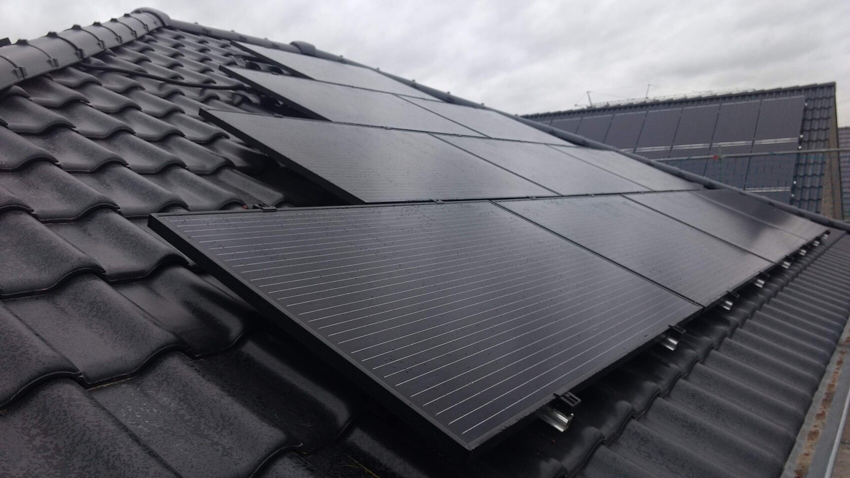 Dunkles Ziegeldach mit mehreren Solarmodulen auf dem Dach unter grauem Himmel.